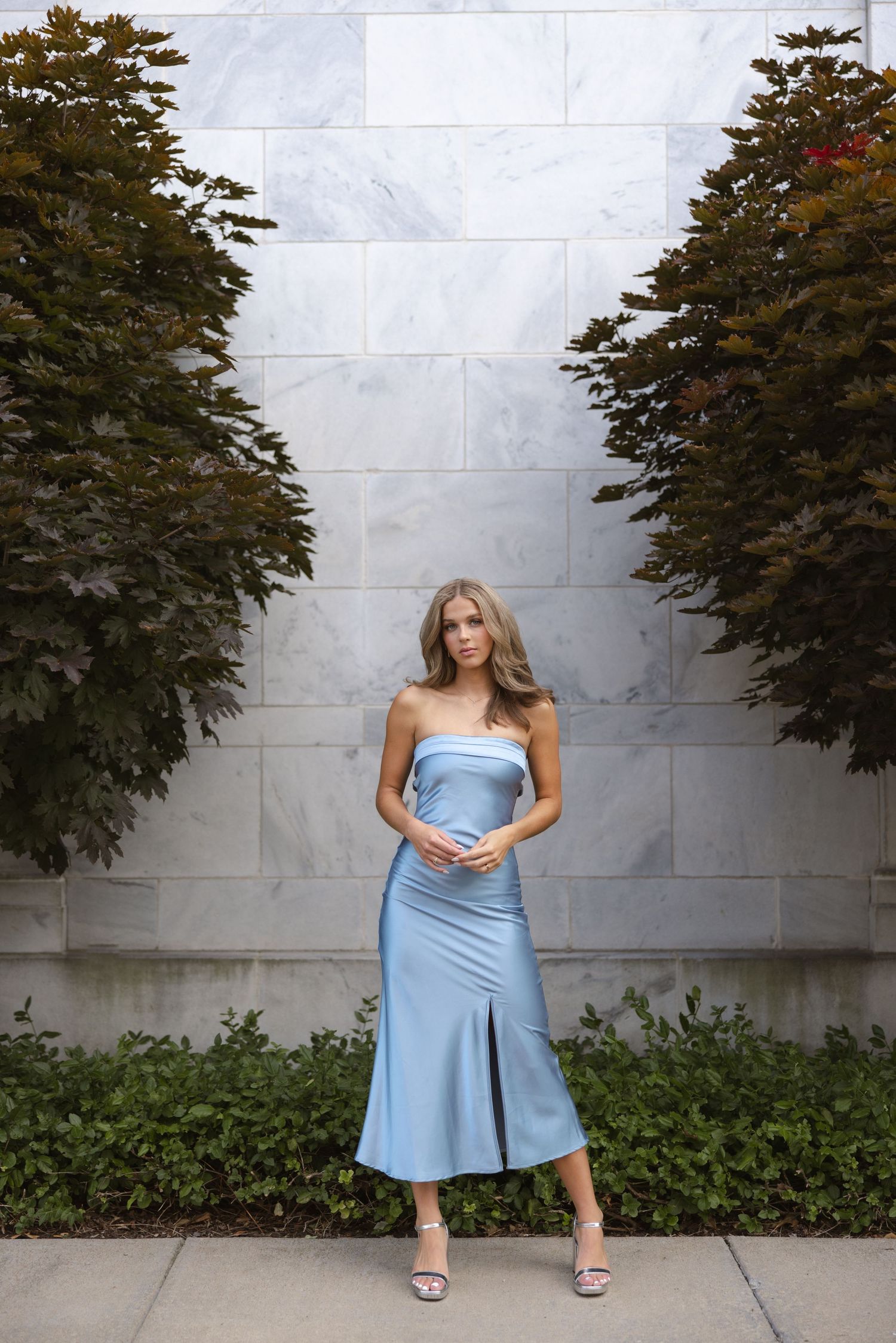 Elegant light blue strapless dress styled against modern stone wall with framing evergreen trees.