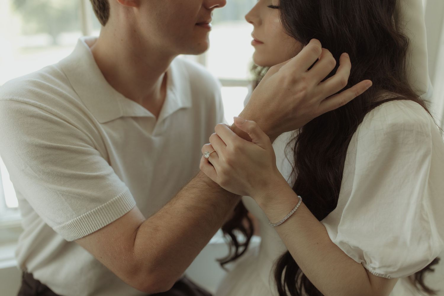 Two people in white clothing share an intimate moment together in soft window light.