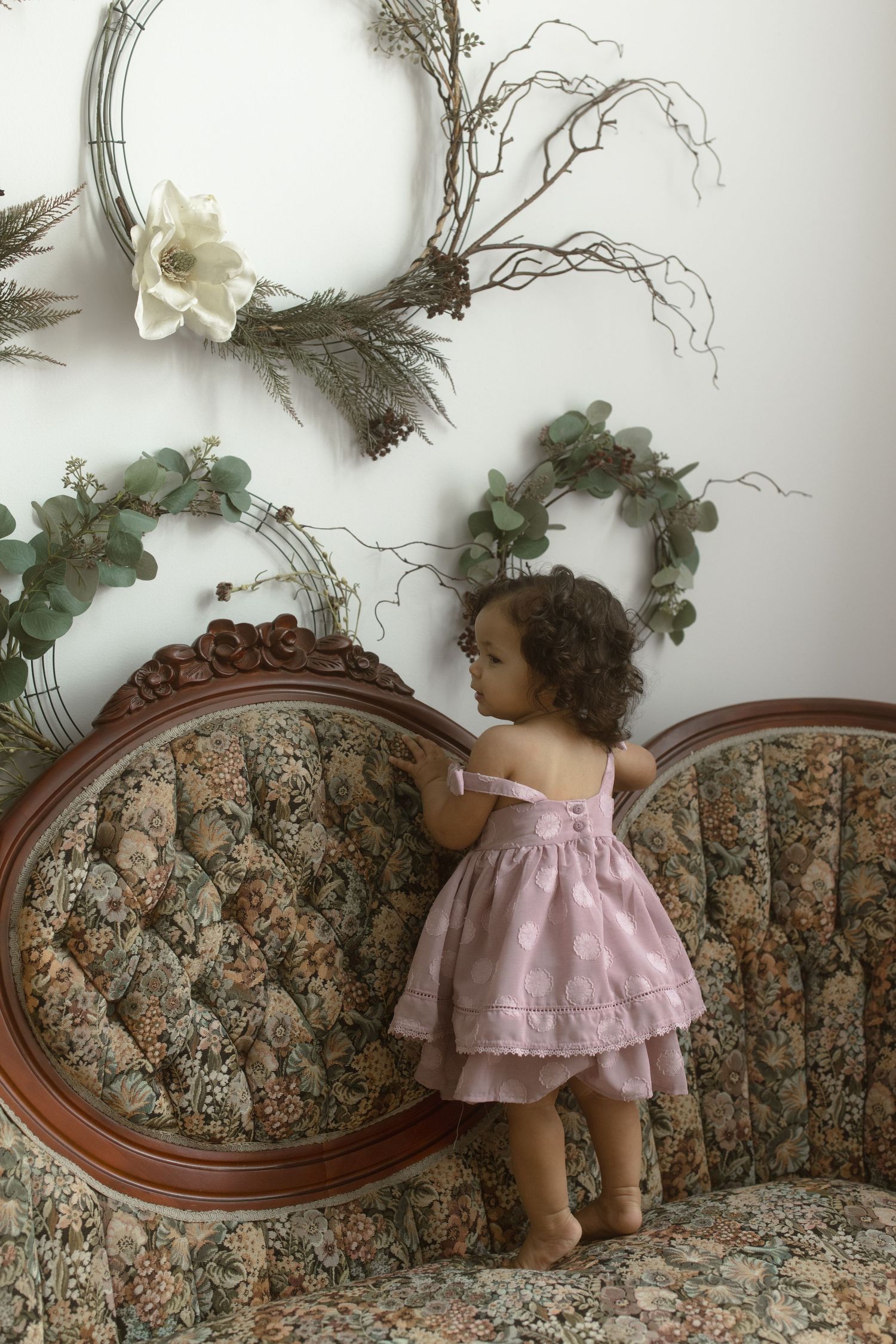 Small child in pink dress standing on vintage floral sofa against white wall with decorative wreath and greenery.