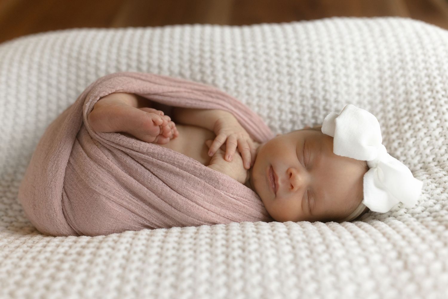 Newborn baby wrapped in soft pink fabric sleeping on white textured blanket with bow accessory.