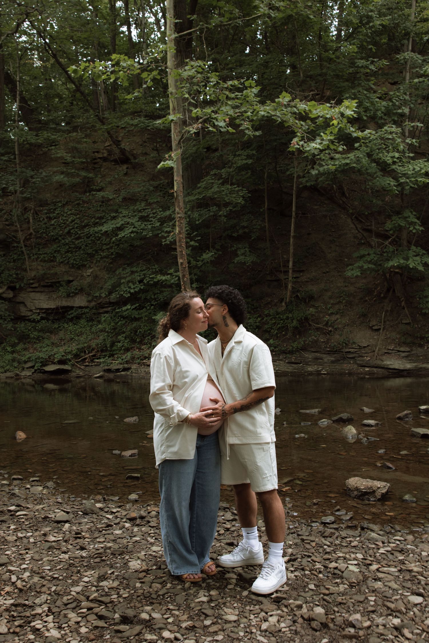 A romantic couple embraces by a creek bed surrounded by lush forest scenery.