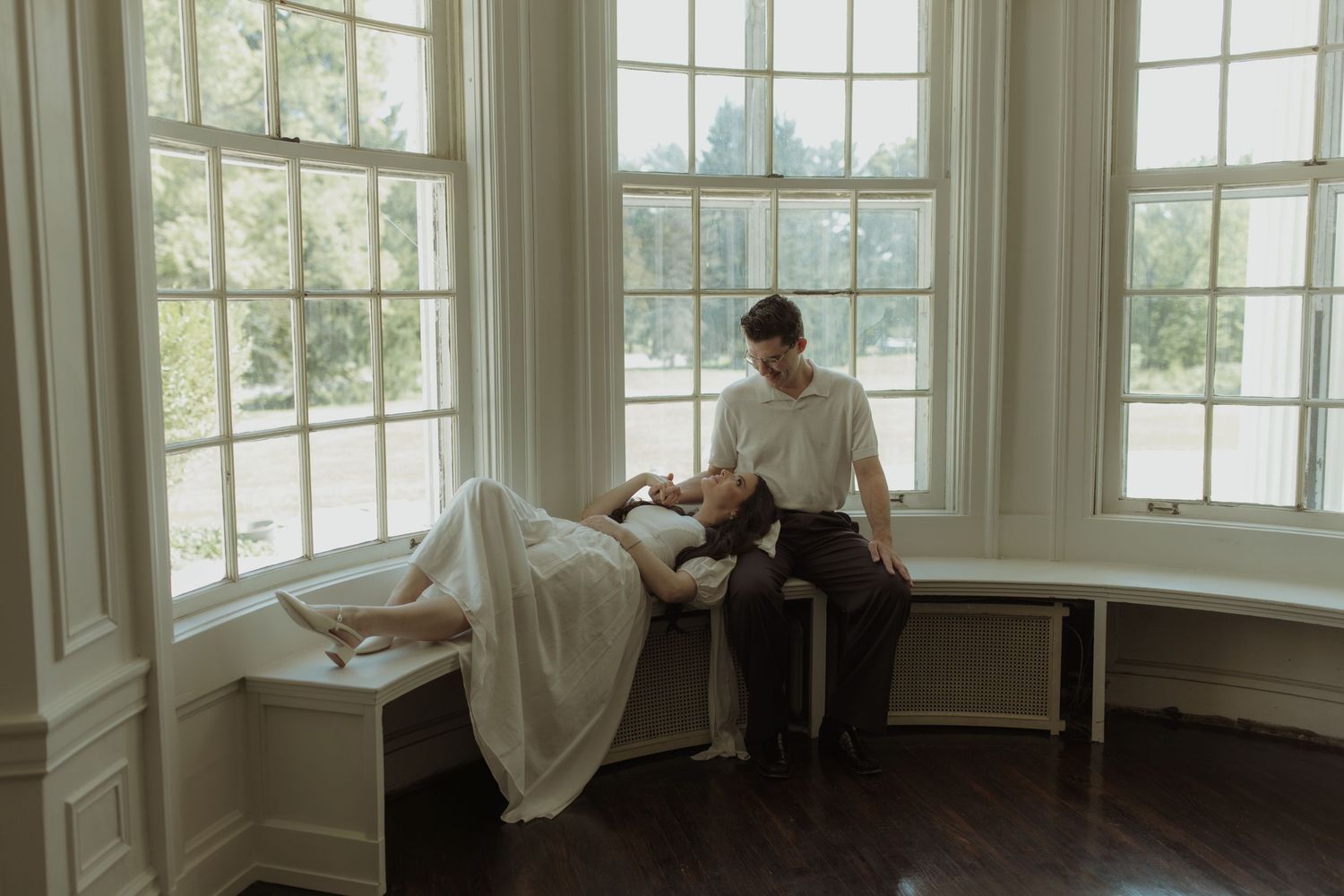 Two figures sit together in a bright bay window alcove with white trim and multiple paned windows.