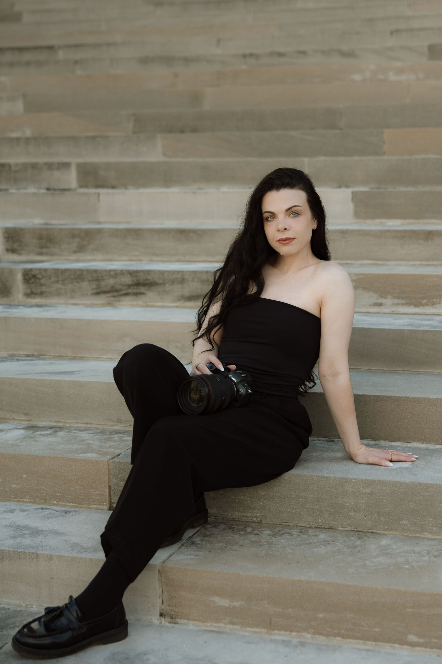 Woman in black strapless jumpsuit sitting on stone steps, holding a camera, with long dark hair flowing to one side.