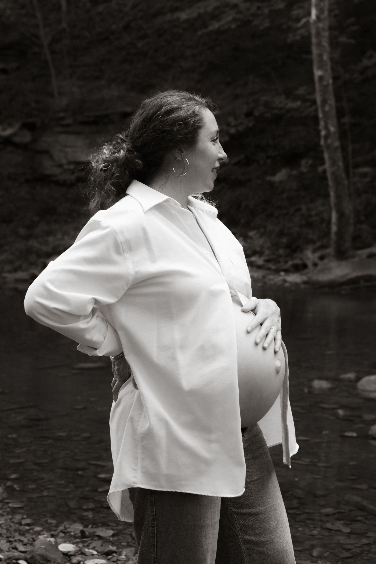 A pregnant person in a white shirt stands in profile against a wooded creek backdrop.