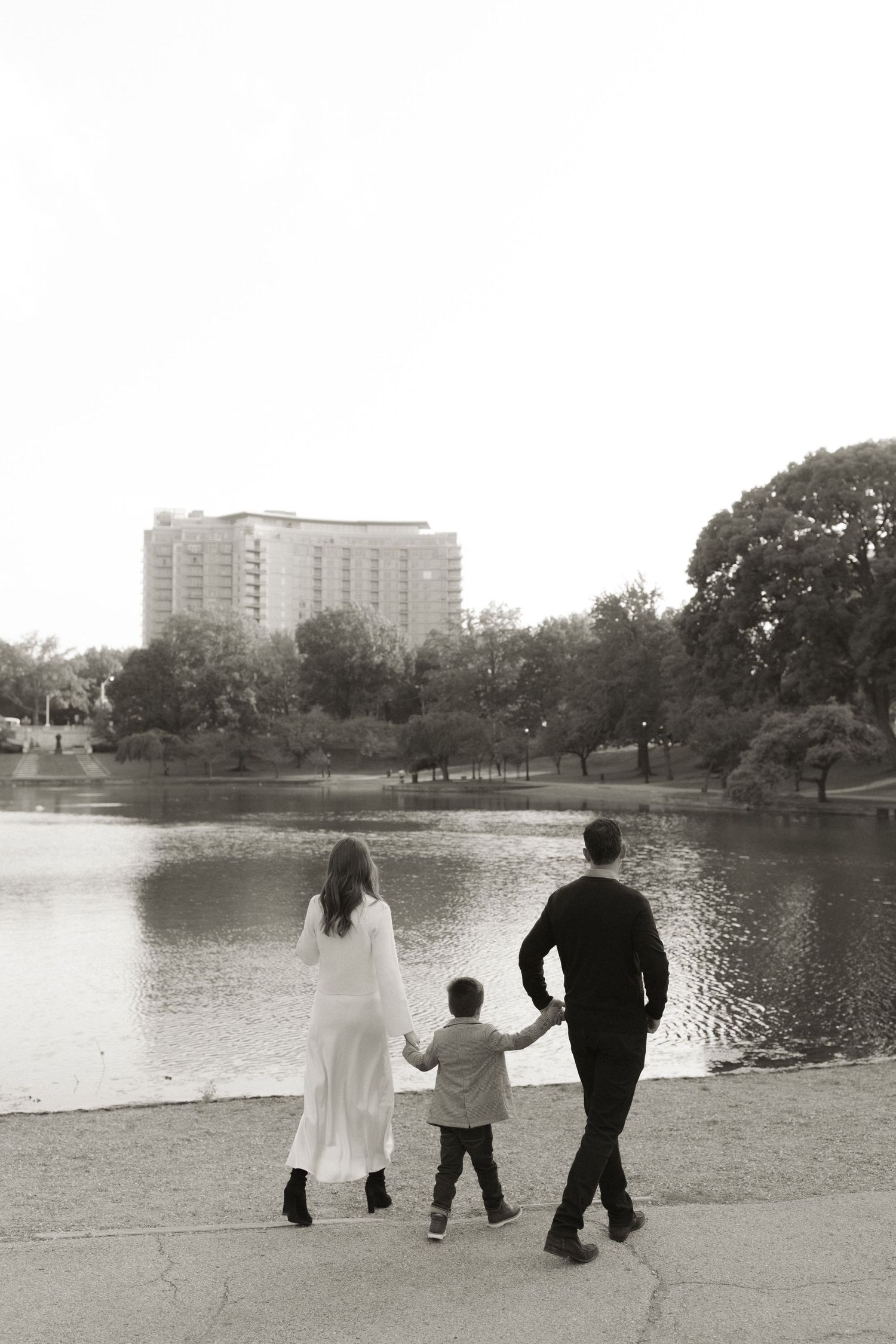 A family holds hands while standing at the edge of a pond with a tall apartment building visible in the distance.