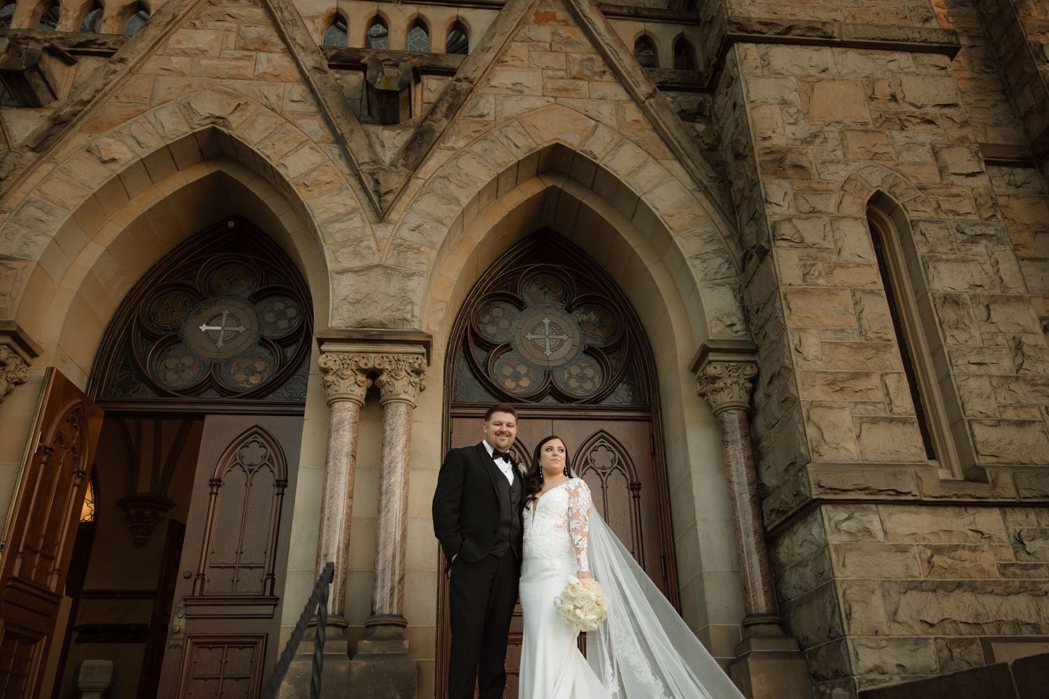 A couple poses under Gothic stone arches at the entrance of a historic church on their wedding day.