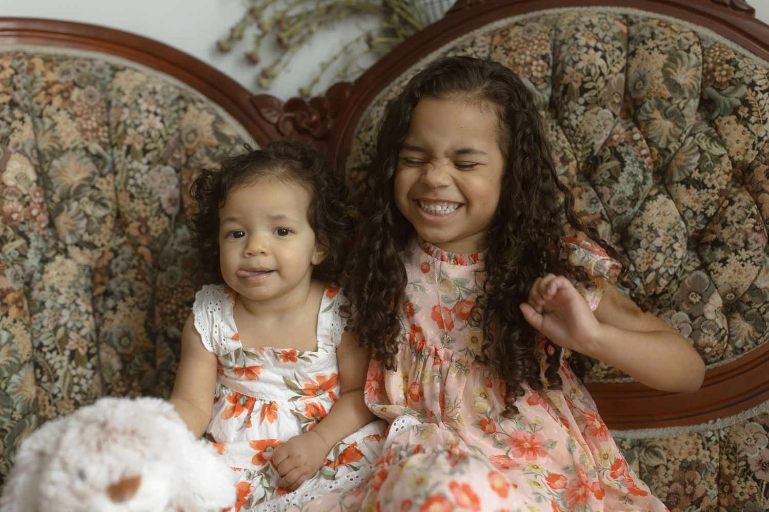 Two young children wearing floral dresses sit and smile together on an ornate vintage sofa with patterned upholstery.