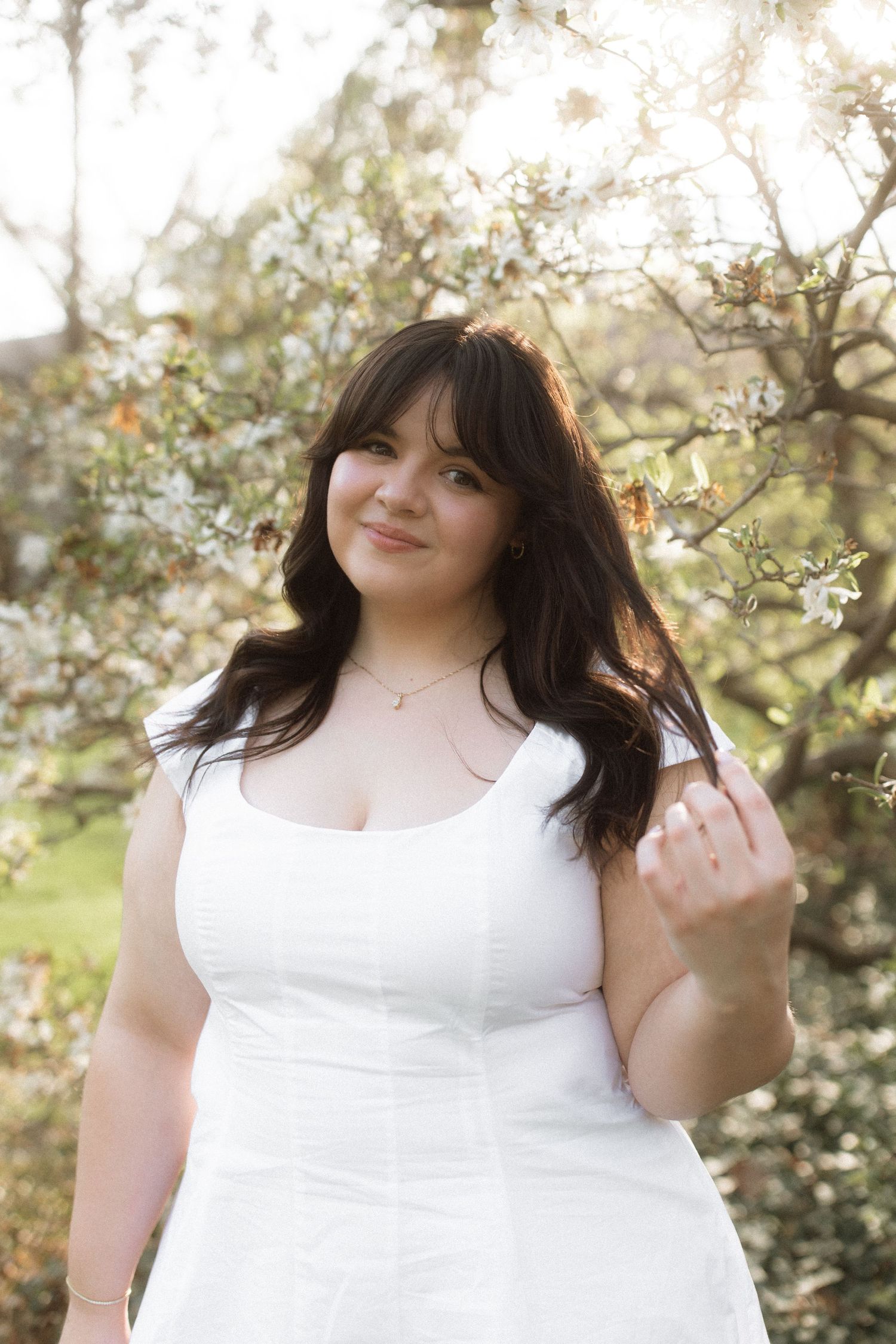 Person in a white sleeveless dress poses outdoors against a backdrop of blooming spring trees and soft natural light.