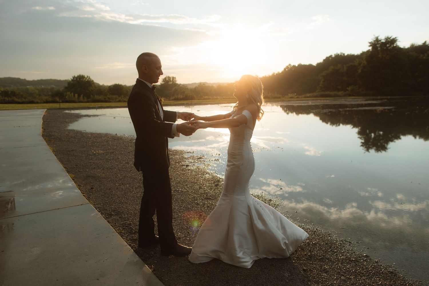 A couple in wedding attire dances by a serene lake at sunset with reflective water and natural scenery.