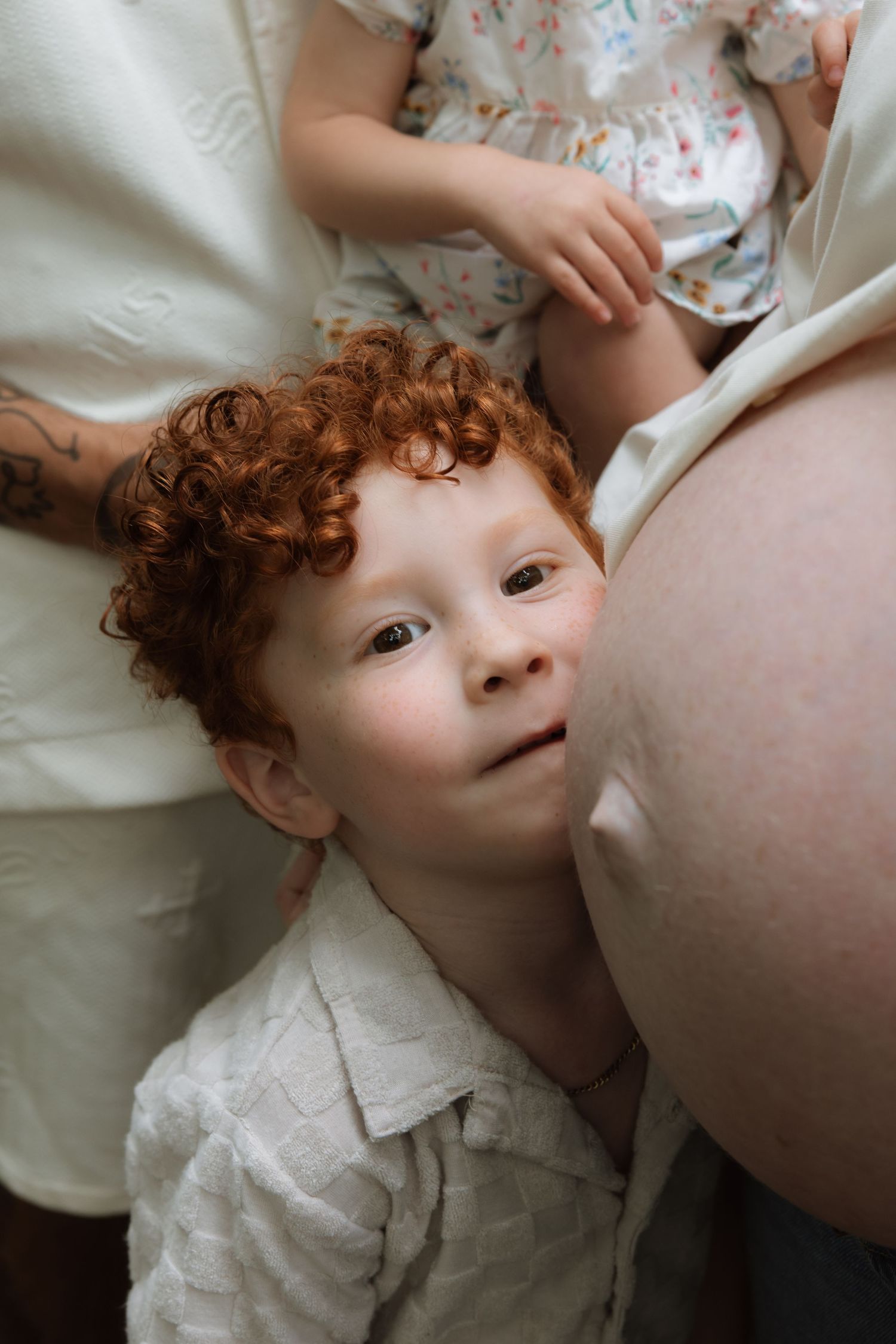 A young child with vibrant red curly hair lies on a white sheet looking up towards someone wearing pajamas.
