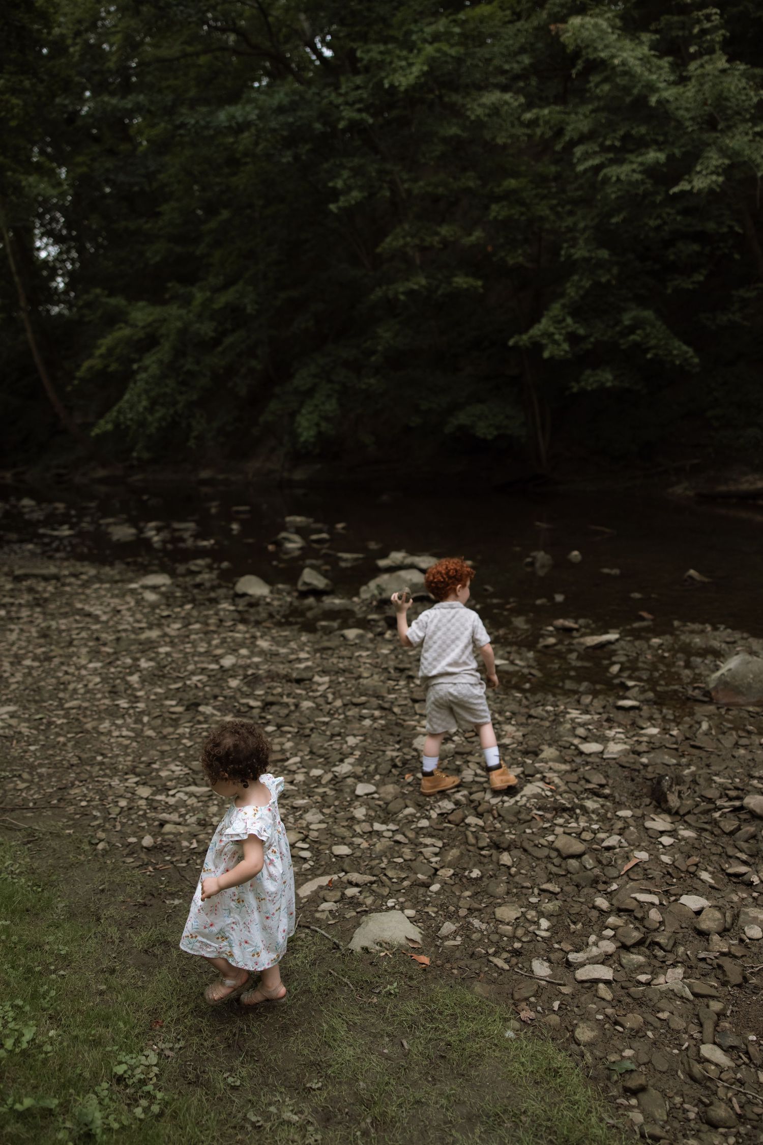 Two children playing along a rocky riverbank surrounded by lush green trees on a summer day.