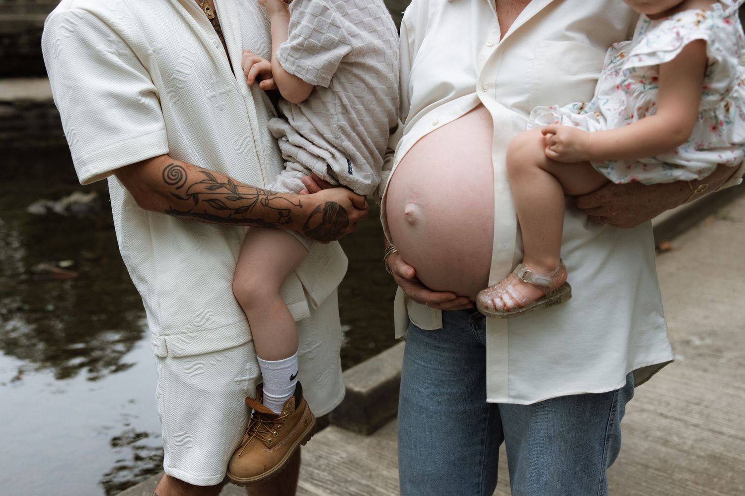 Two people share a tender moment while showing off a pregnant belly near a waterfront.