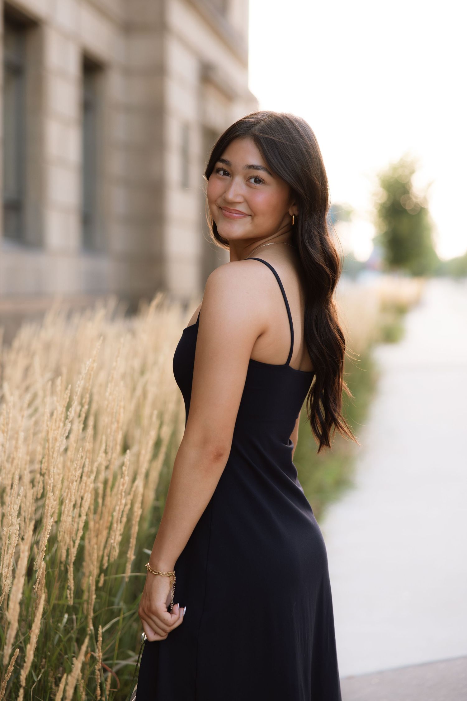 A person in a black dress poses outdoors against a backdrop of tall wheat grass and sunlit architecture.