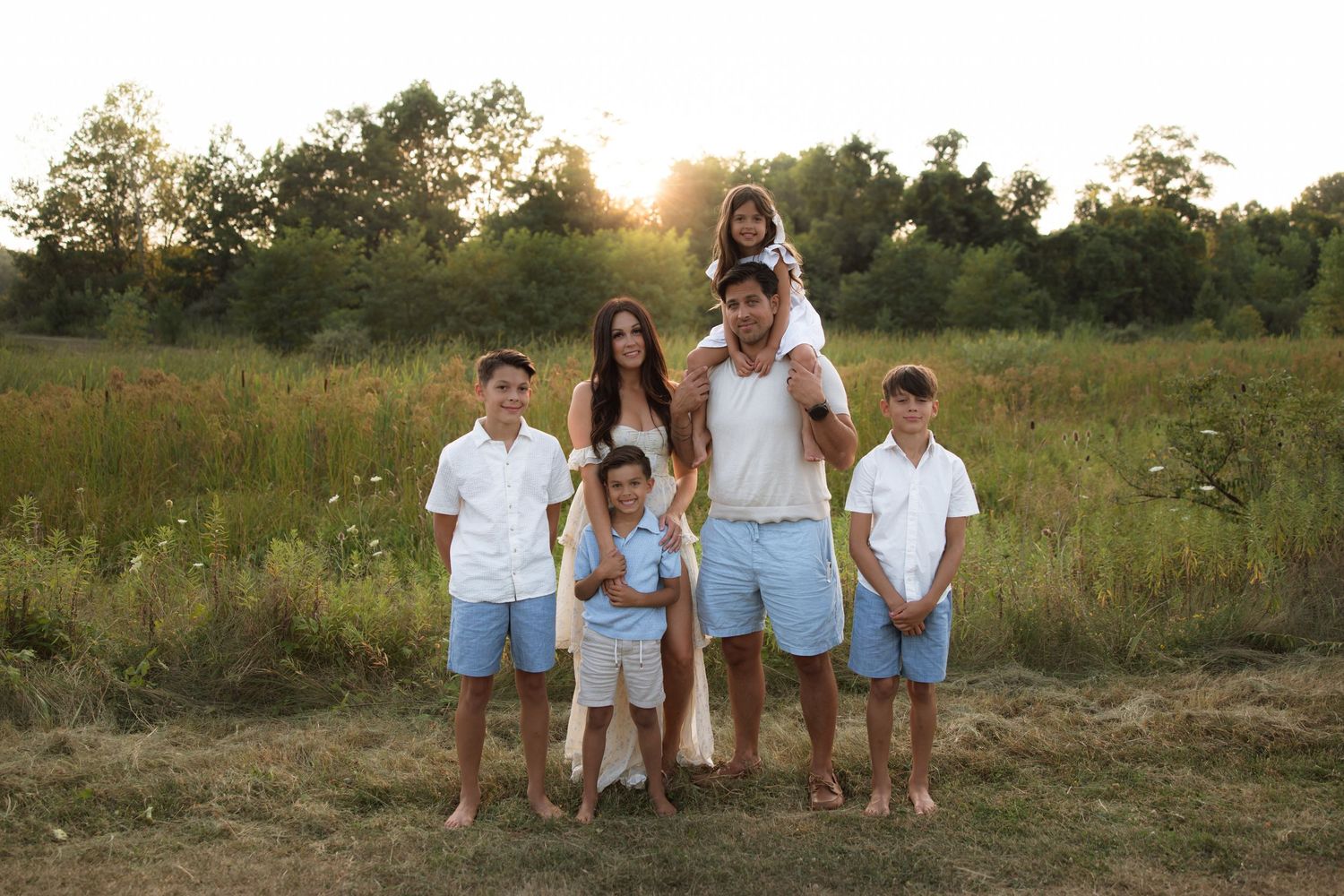A group stands together in coordinating white and blue outfits during a sunset photoshoot in a rustic outdoor field.