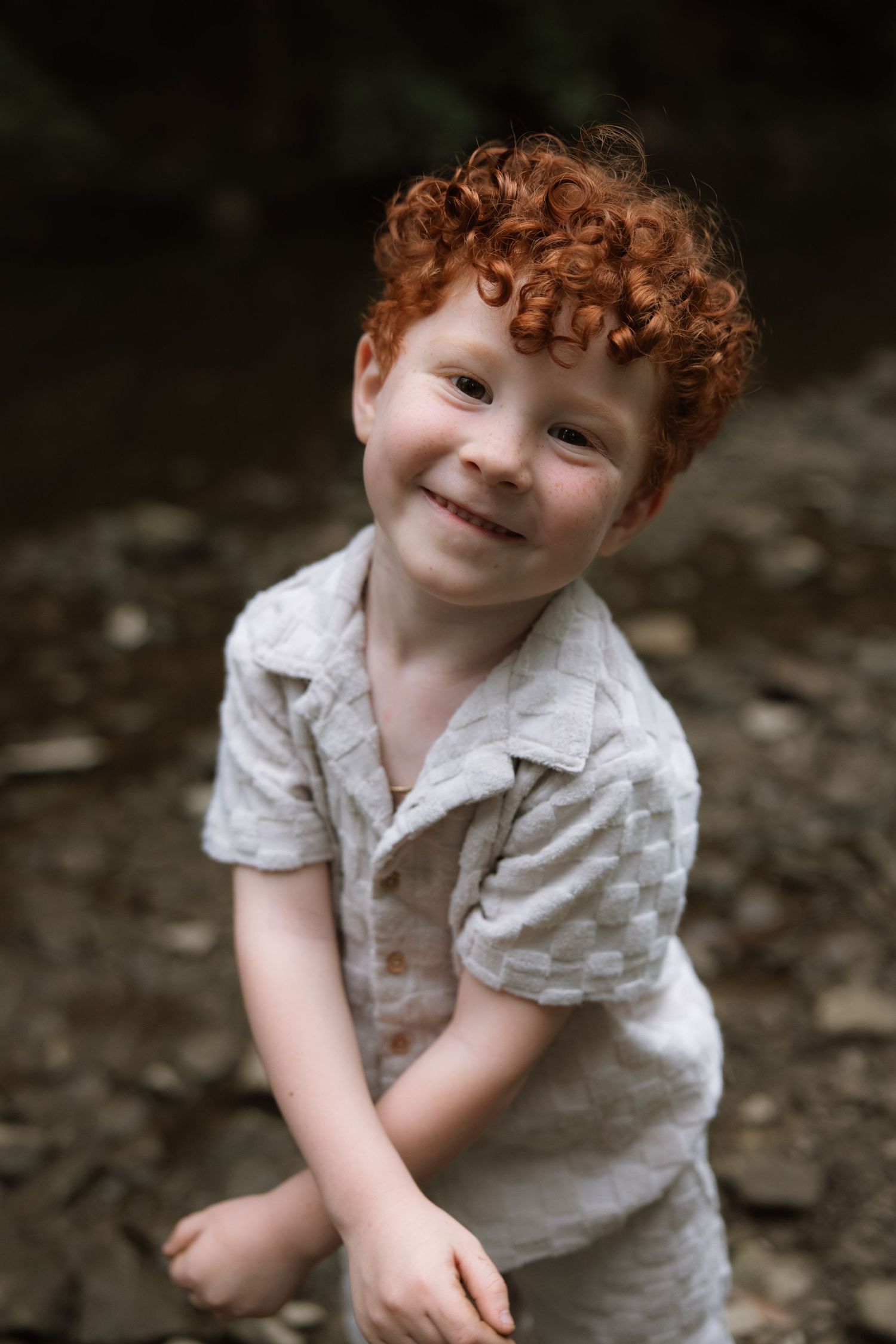 Playful young child with bright curly red hair wearing light colored shirt outdoors.