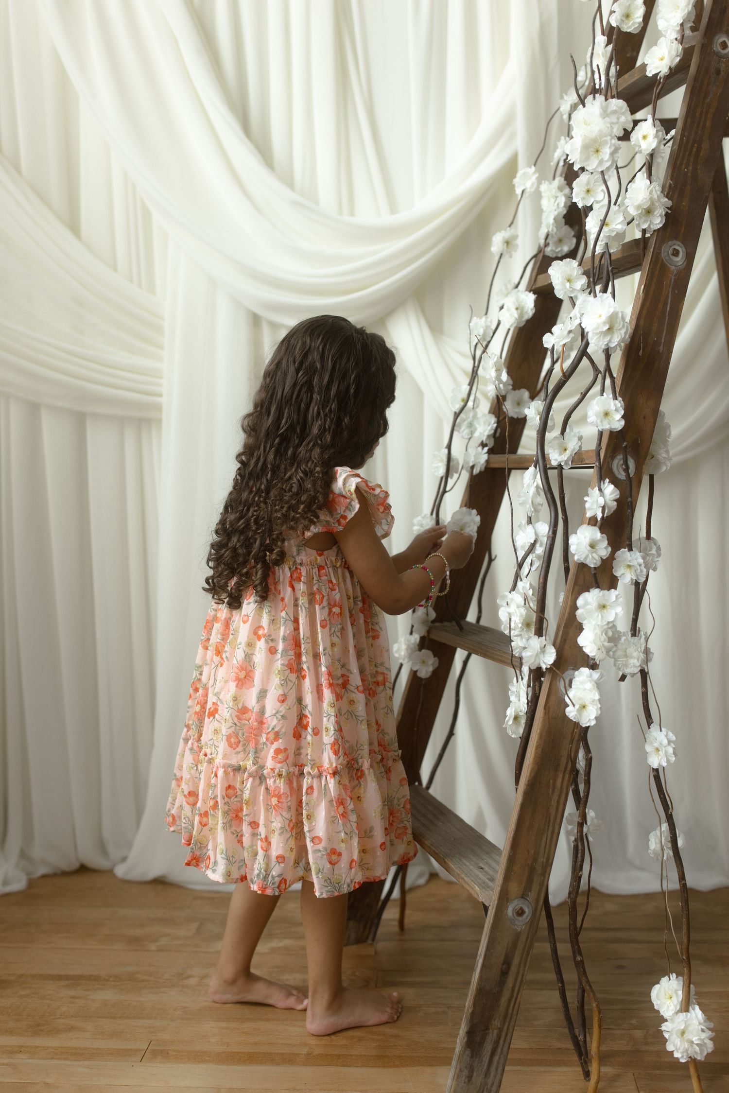 A small child in a floral dress decorates a rustic ladder with white cotton branches against draped fabric backdrop.