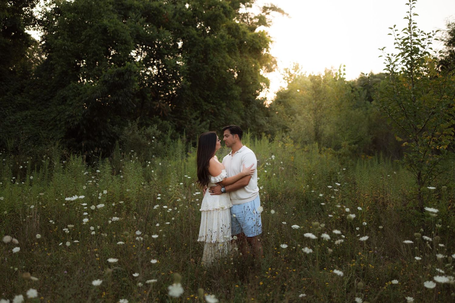A couple in white and blue outfits embrace tenderly in a sunlit meadow filled with wildflowers at sunset.