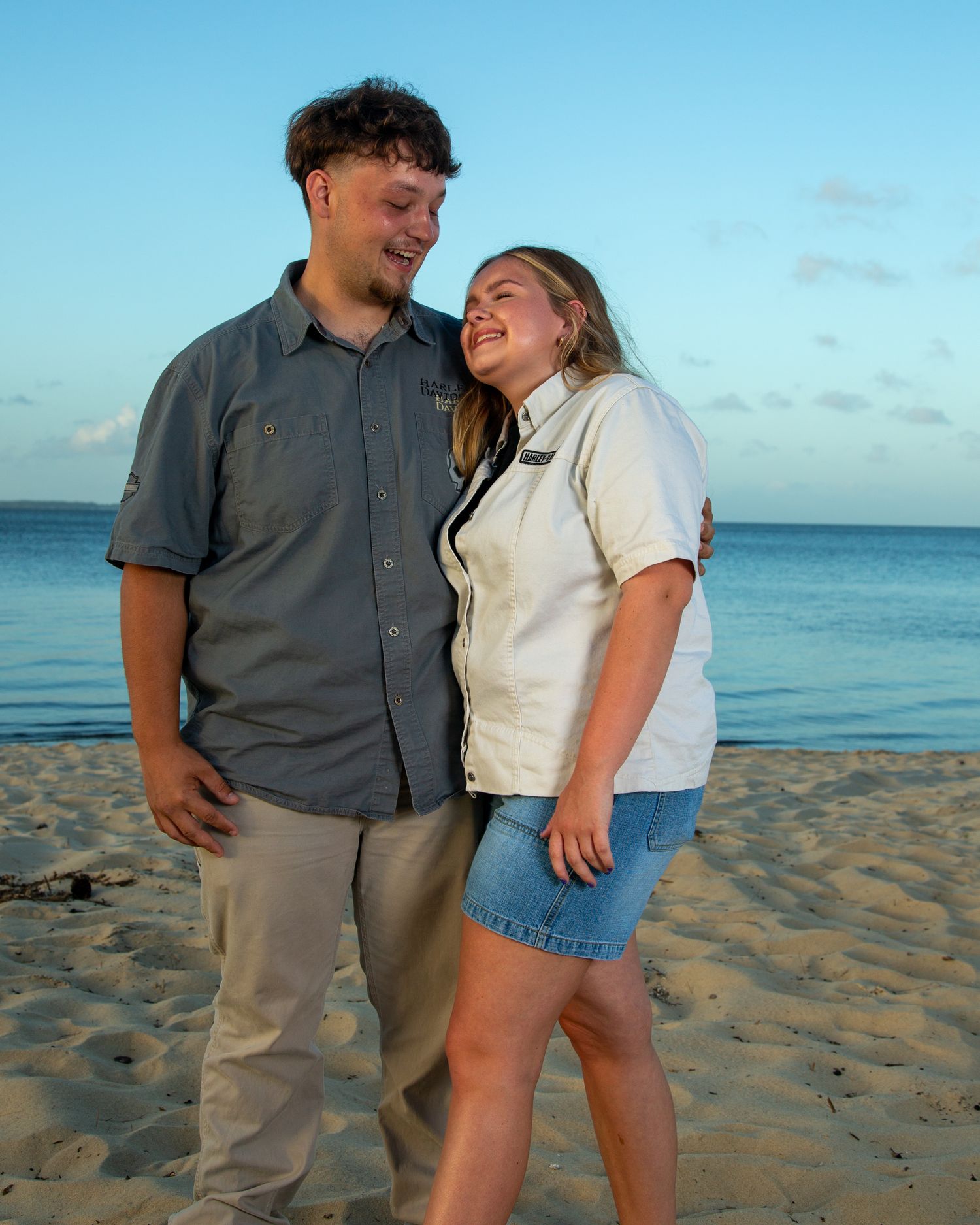 A couple smiles and embraces on a sandy beach at sunset with calm blue ocean waters in the background.