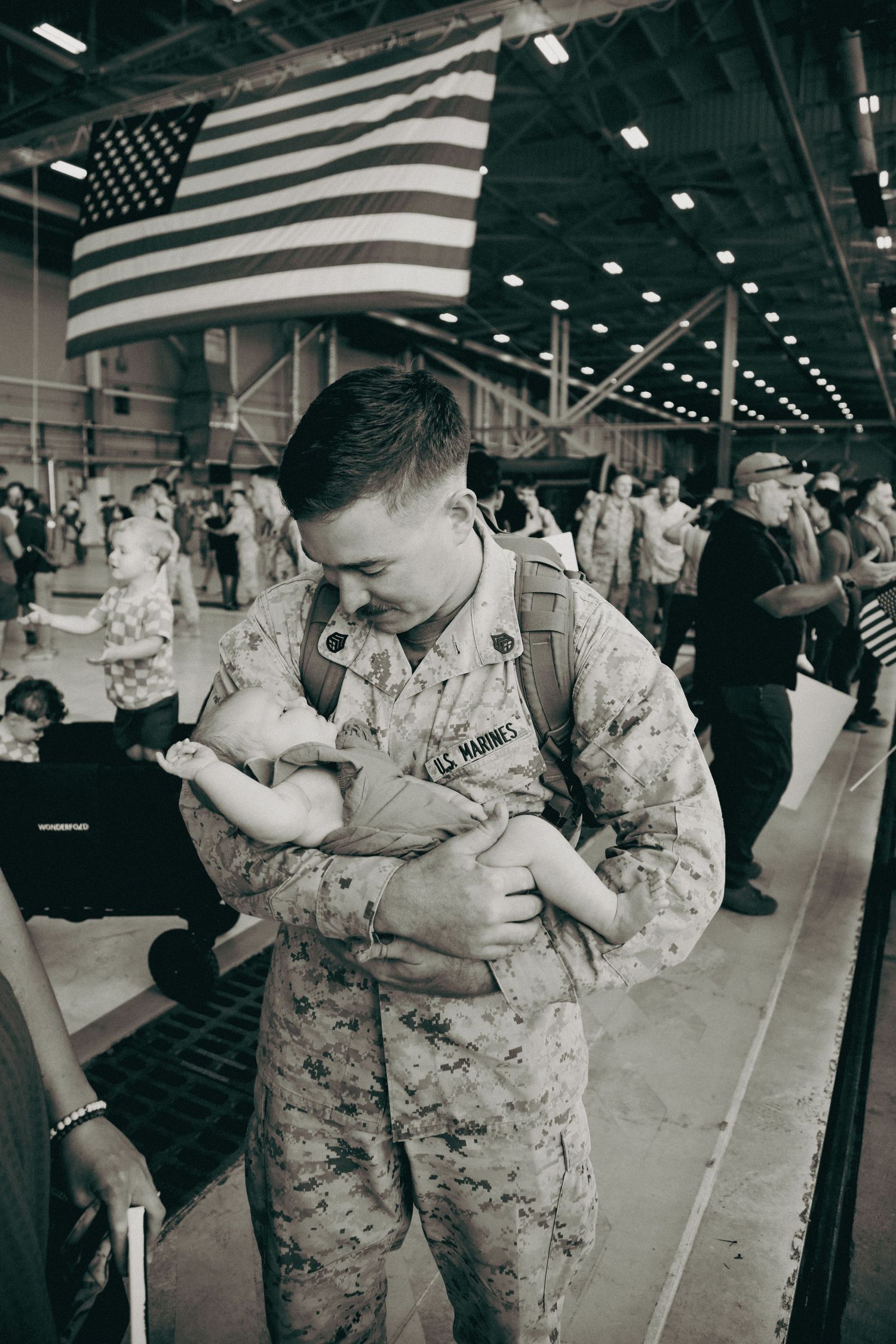 Military service members in camouflage uniforms embrace during an emotional reunion under an American flag.