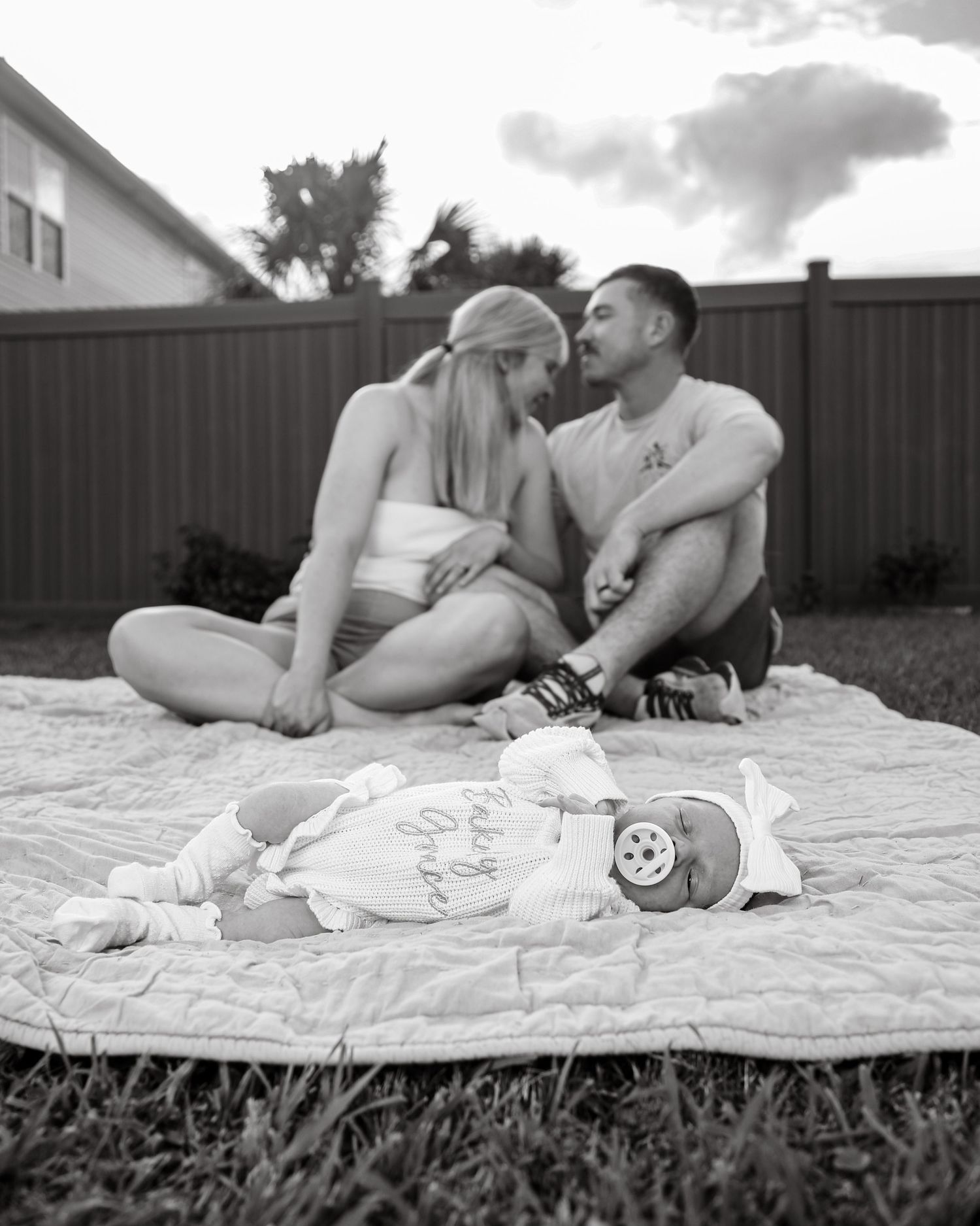 A couple sits together on a blanket outdoors with baby items laid out next to them in black and white photography.