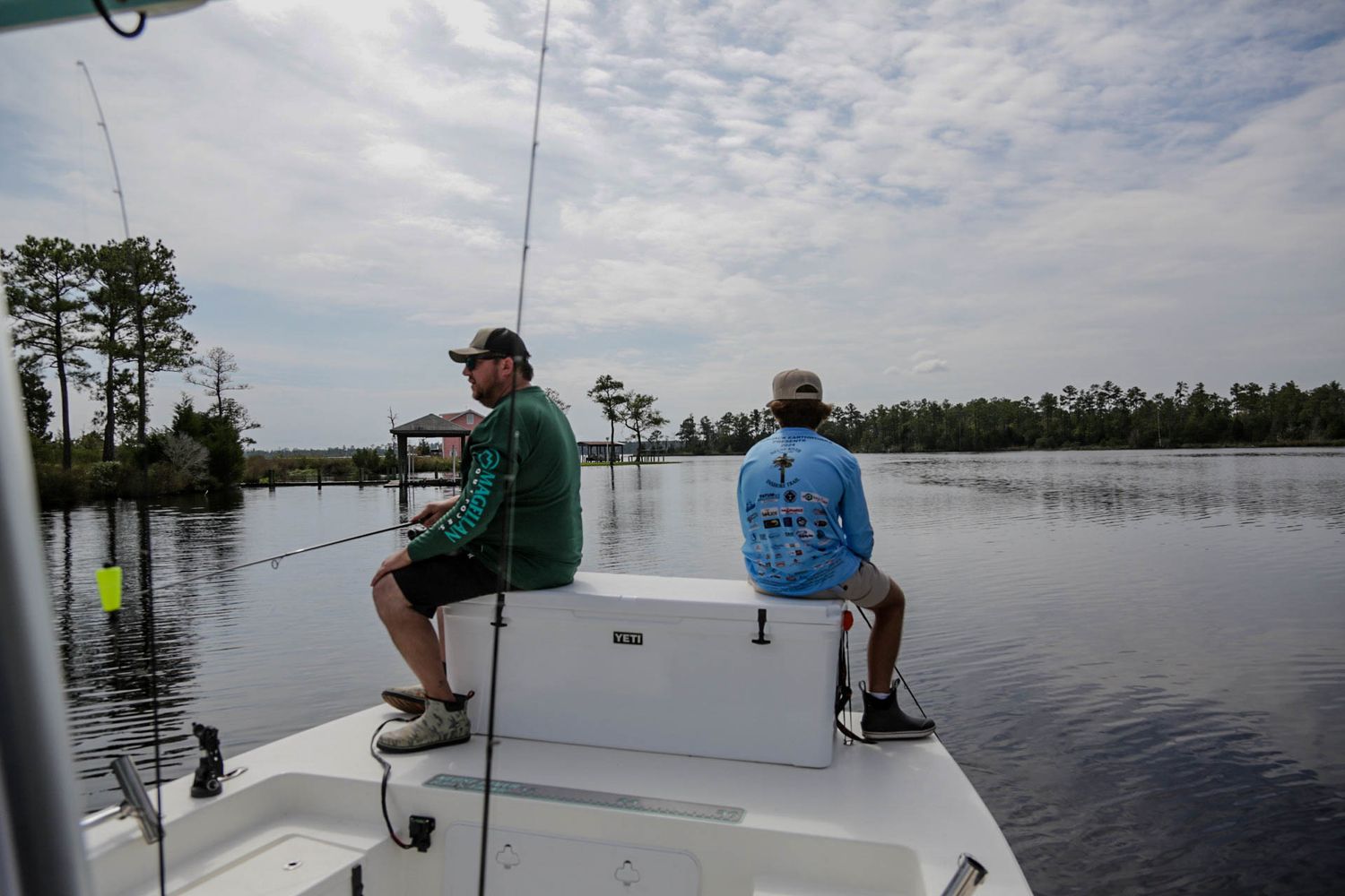 Two anglers fishing from the bow of a boat on a tranquil lake surrounded by pine trees.