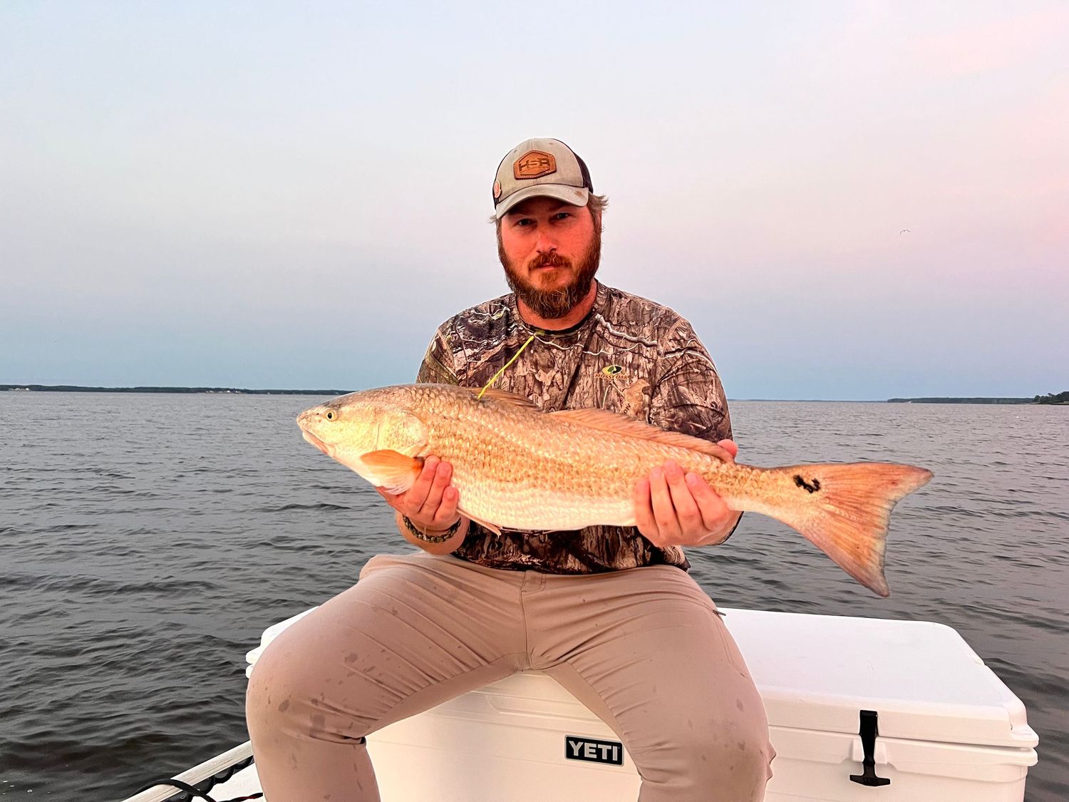 Angler holding large redfish at sunset with YETI cooler visible.