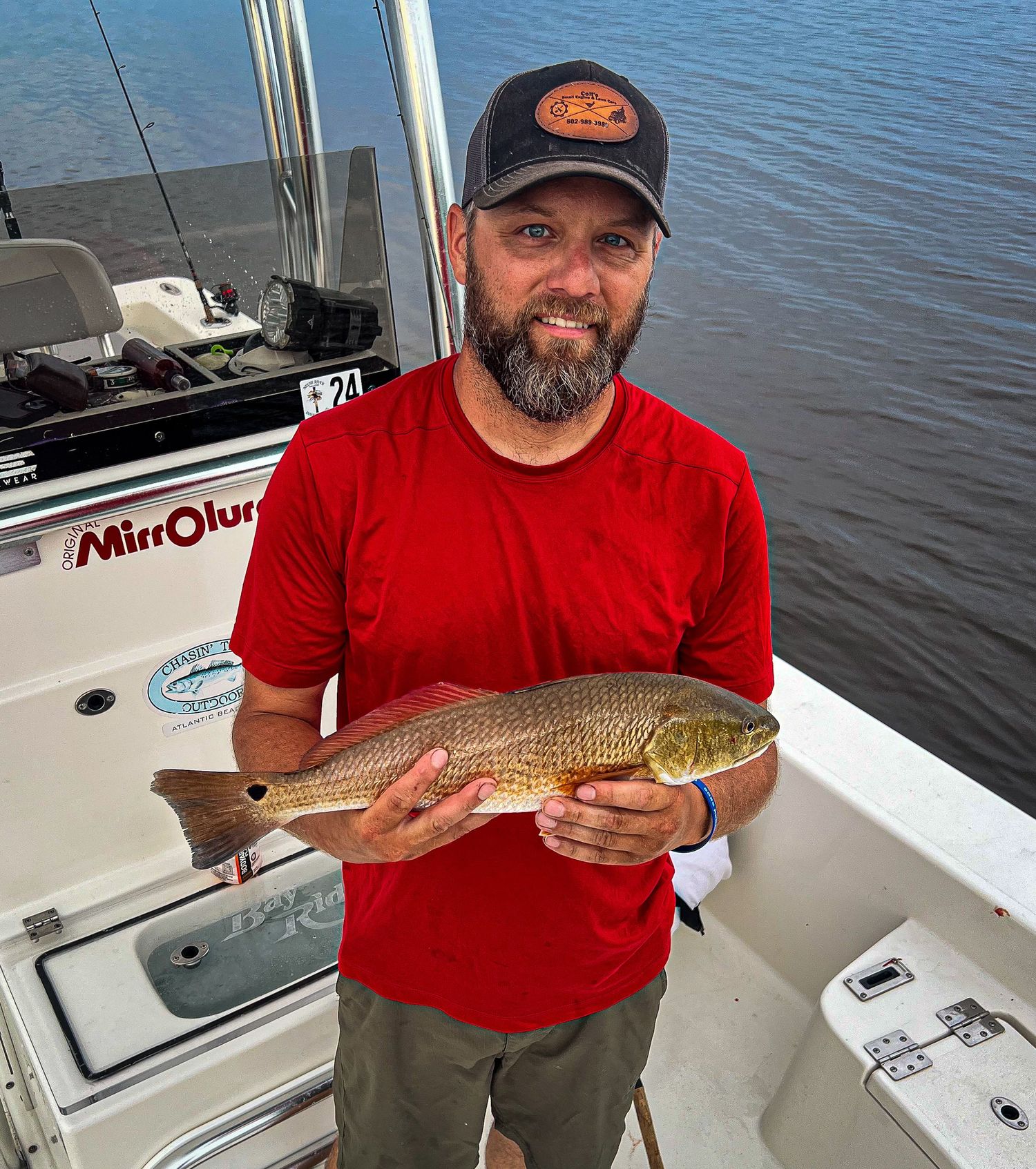 Fisherman wearing red shirt displays a fresh caught redfish from boat on calm waters.