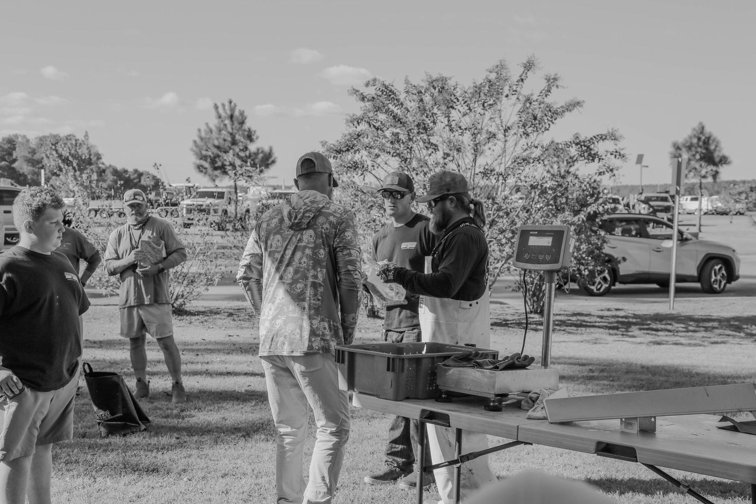 Group gathers around outdoor table with fishing equipment and supplies in park setting.