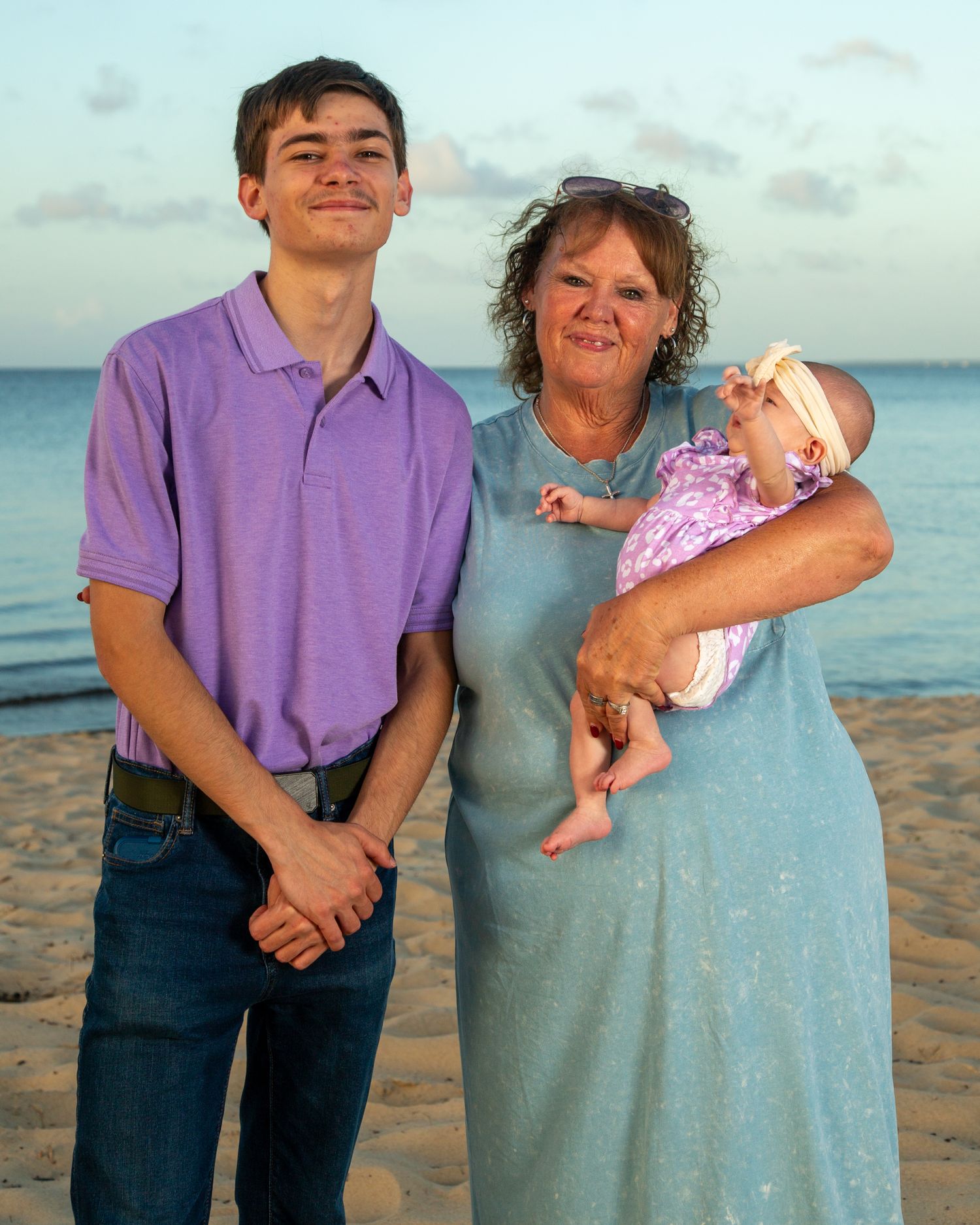A teenage boy and older woman holding a newborn baby pose together on a sandy beach at sunset.