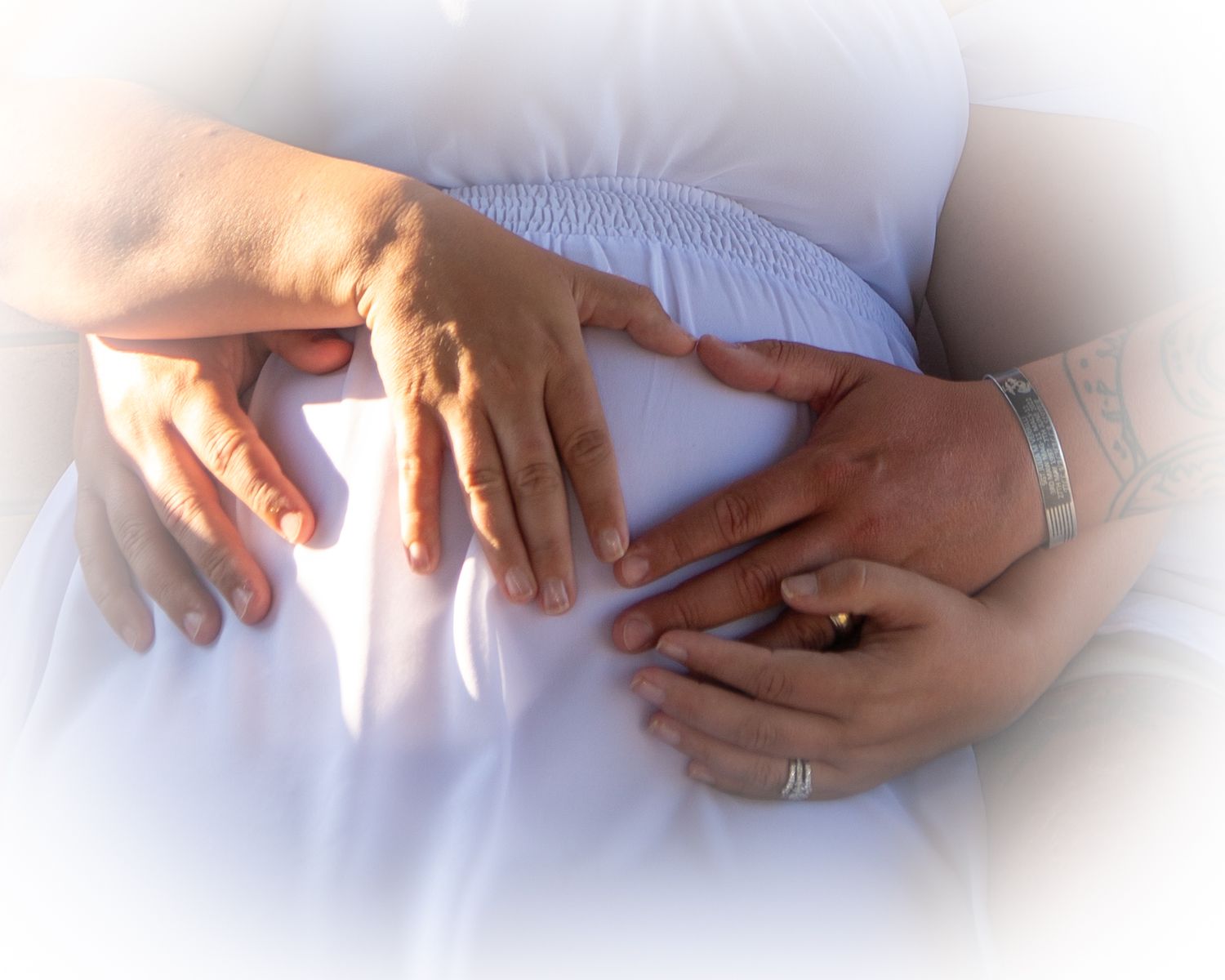 Couple tenderly holding pregnant belly with hands, wearing rings, close-up maternity photo in soft white light.