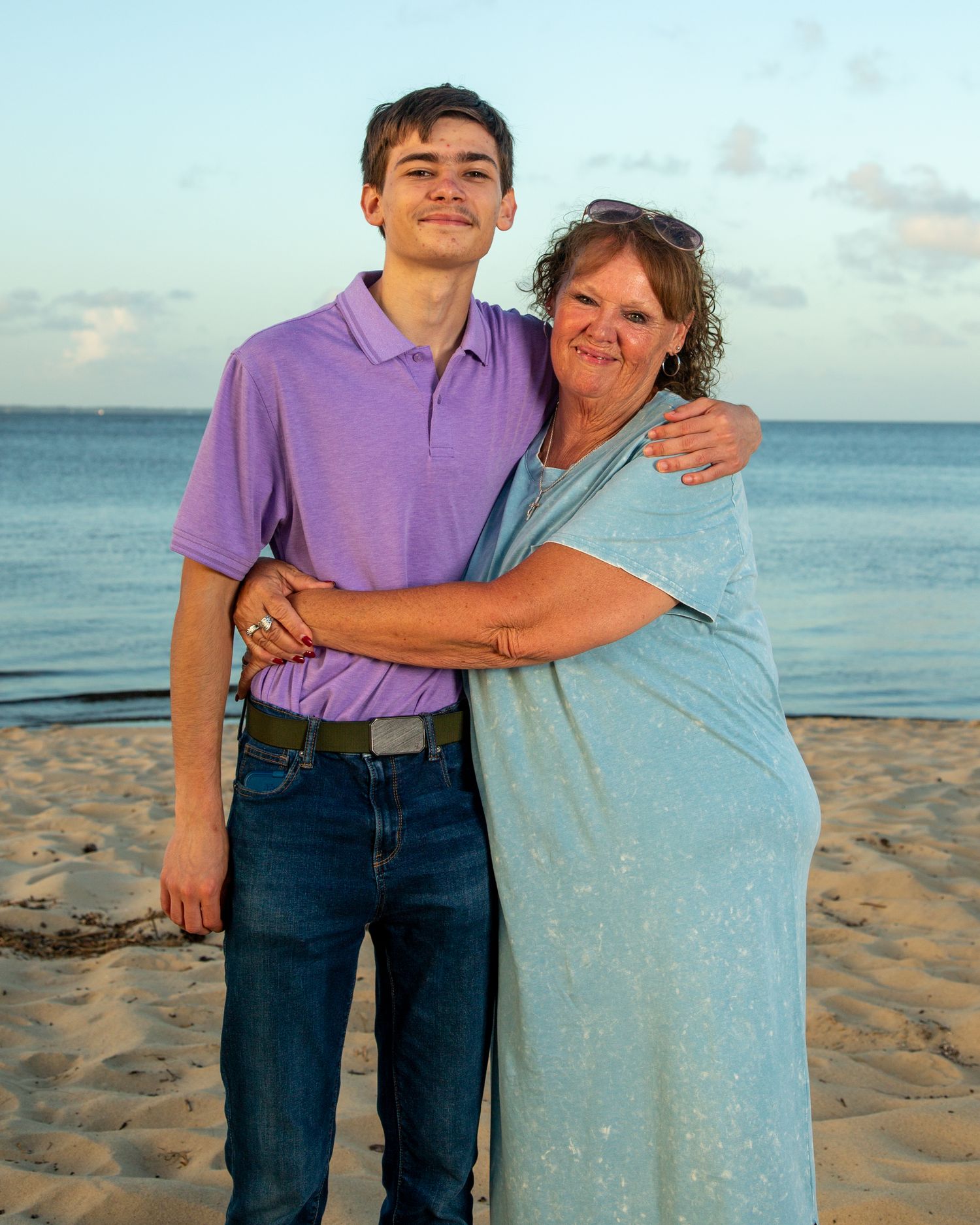 A teenage boy and older woman embrace warmly on a sandy beach at sunset, wearing casual summer attire.