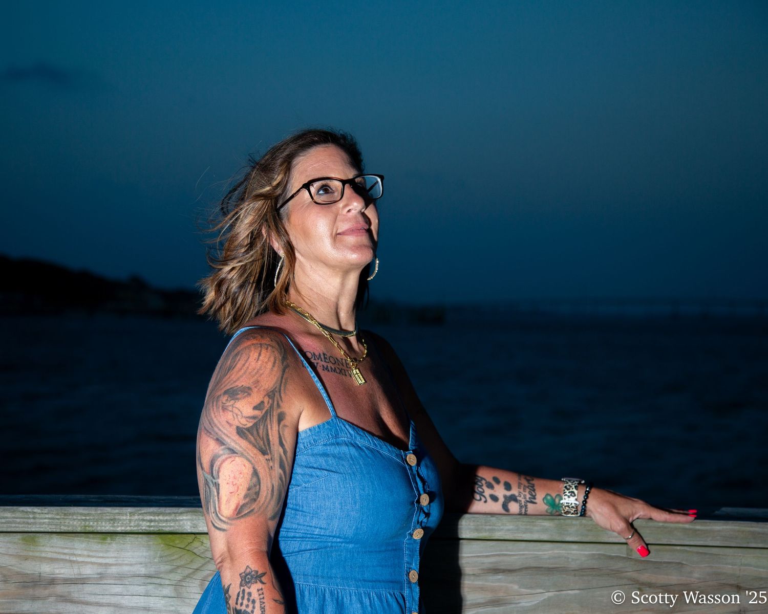 Tattooed woman in blue dress wearing glasses poses on a wooden dock at dusk by the ocean.
