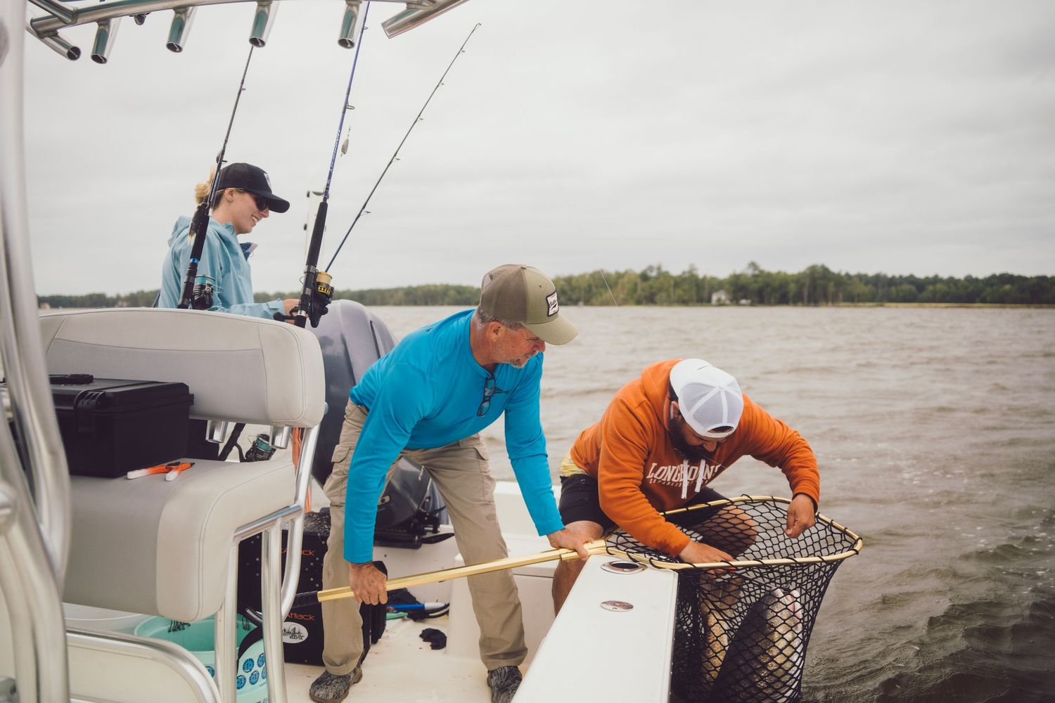 Three anglers on a fishing boat hauling in a catch with a net on a cloudy day near a tree-lined shore.