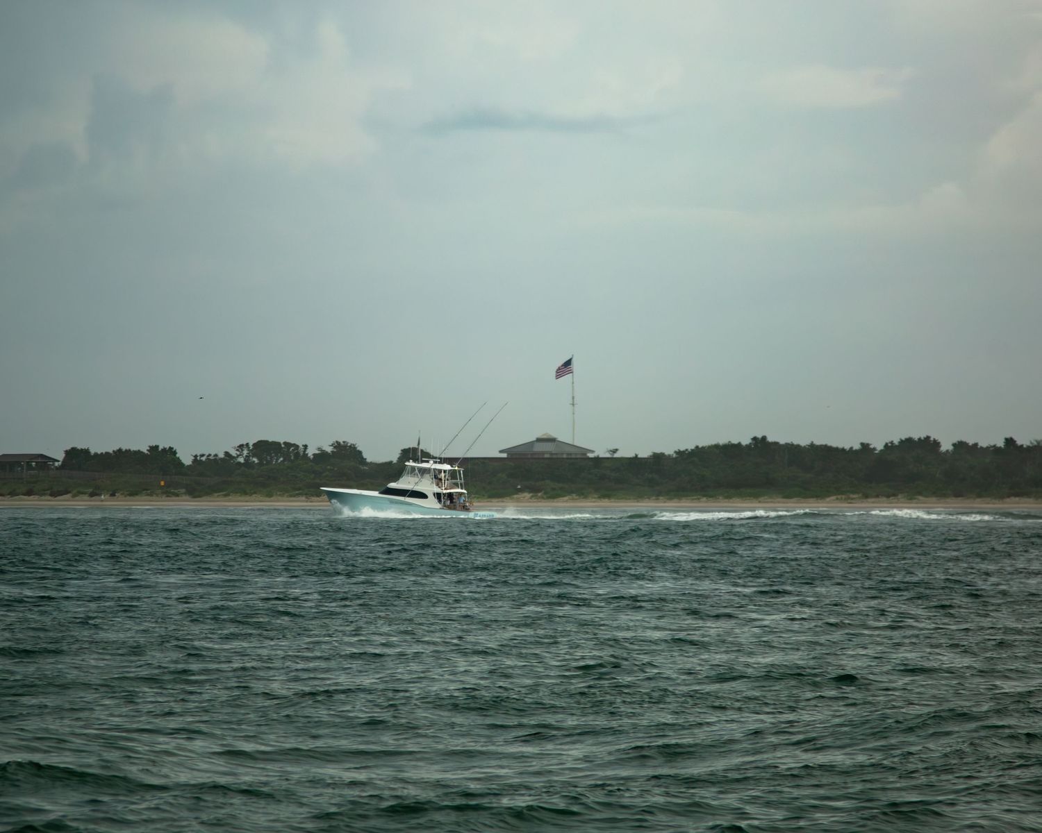 A white sport fishing boat cruises along the coastline with a flag waving in the background.