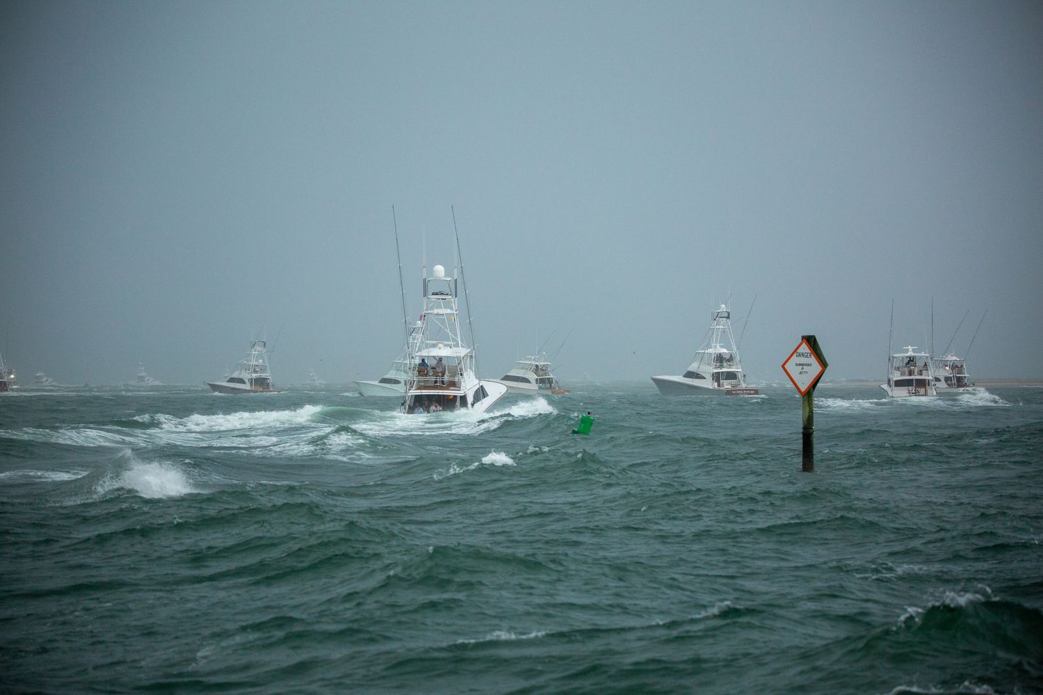 Fleet of sport fishing boats navigating rough waters around channel markers in stormy conditions.