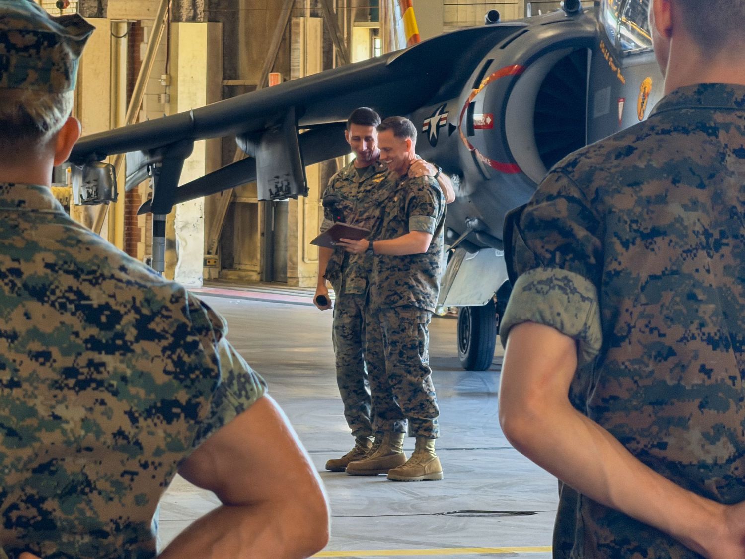 Two Marines in camouflage uniforms embrace in a military hangar near a fighter jet, surrounded by fellow service members.