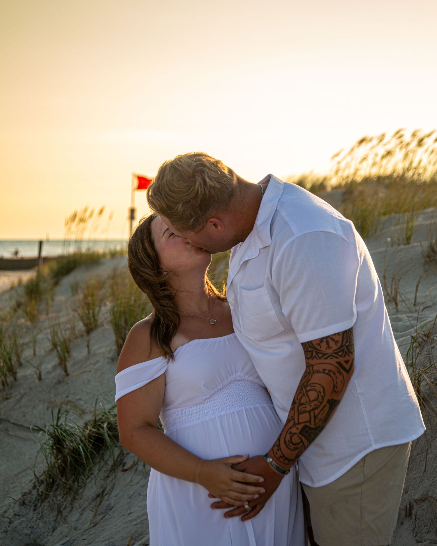 Couple kissing on sandy beach dunes at golden sunset, woman in white off-shoulder maternity dress, man with tattoo sleeve.