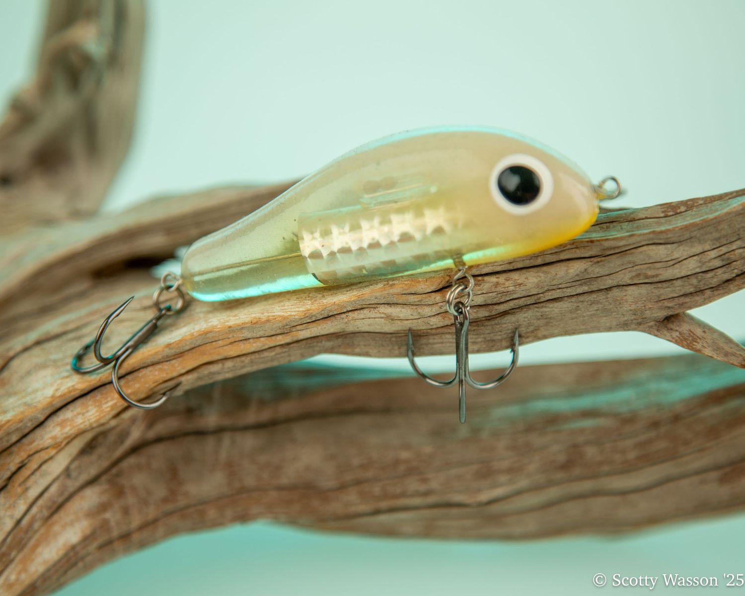 A shiny green and gold fishing lure with treble hooks resting on a piece of driftwood against a teal background.