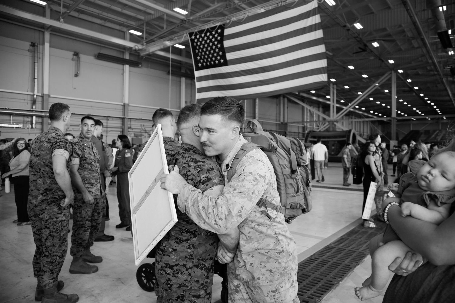 Military personnel reunite with loved ones during an emotional homecoming event under American flags in a large hangar.