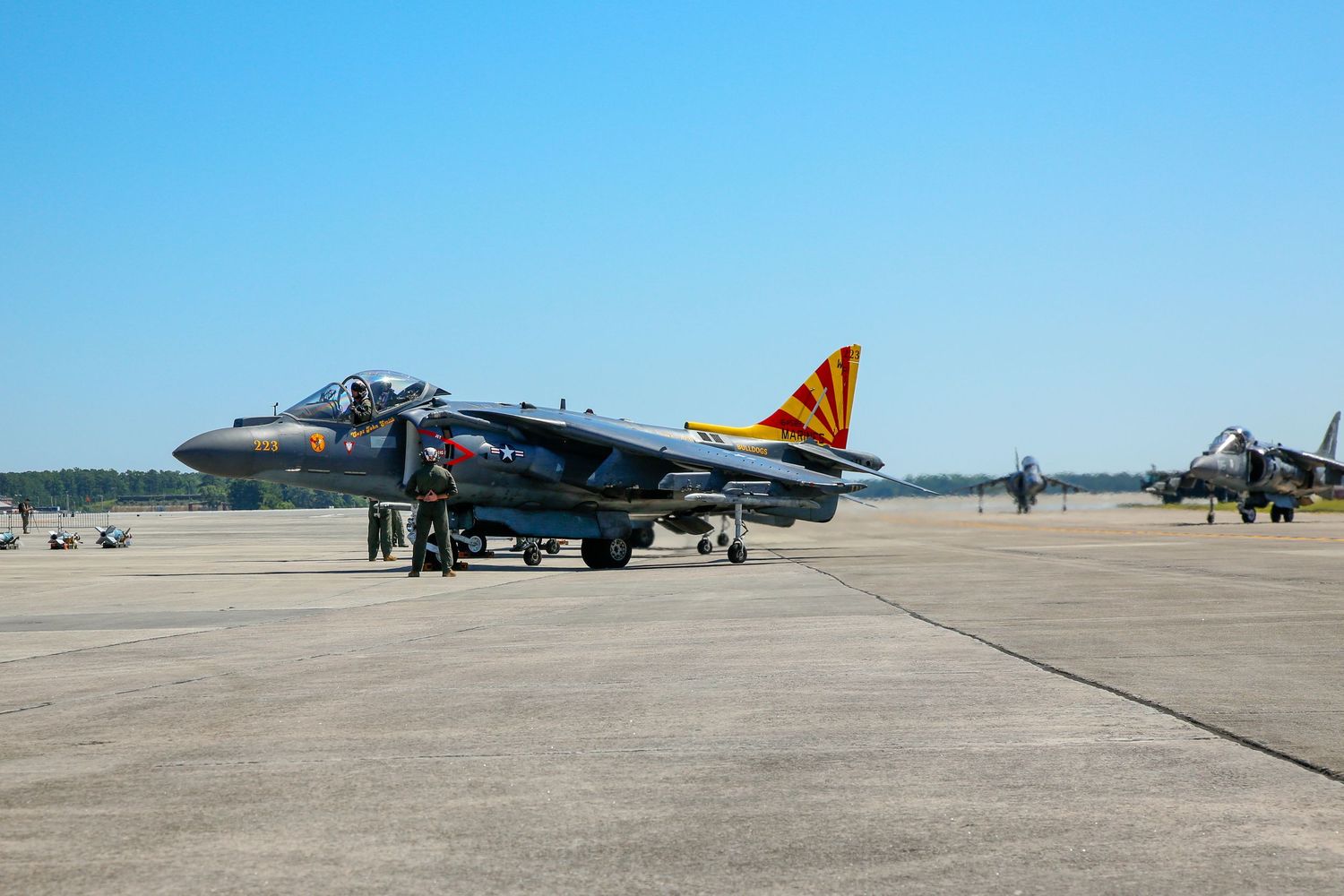 AV-8B Harrier jet aircraft on tarmac with colorful tail markings at military air base on clear day.