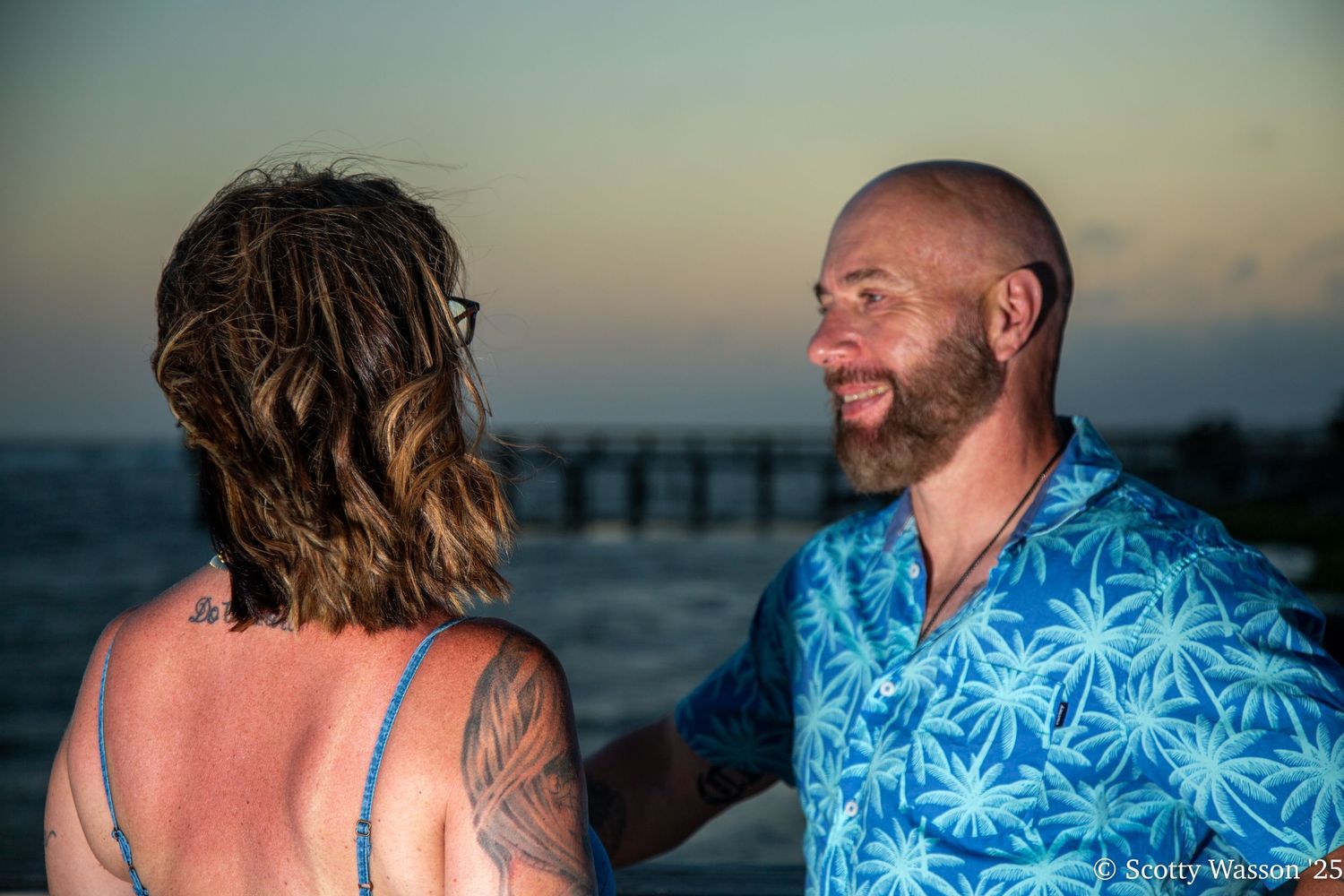 Couple sharing a romantic moment on a beach at sunset, man in blue floral shirt smiling at woman with tattoos near a pier.