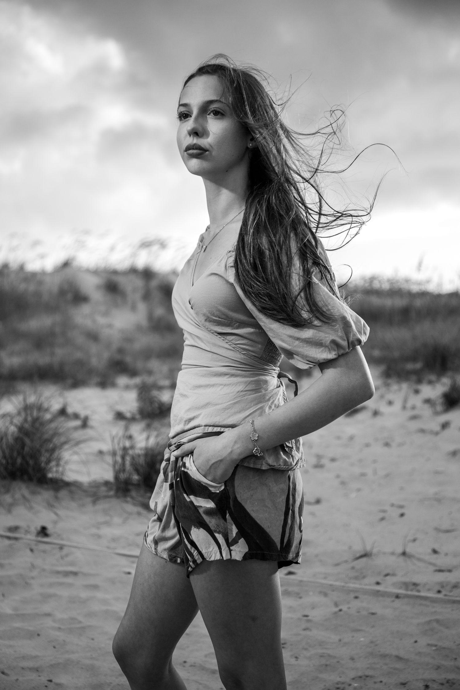 A dramatic black and white portrait of a young woman with windswept hair standing on a sandy beach.