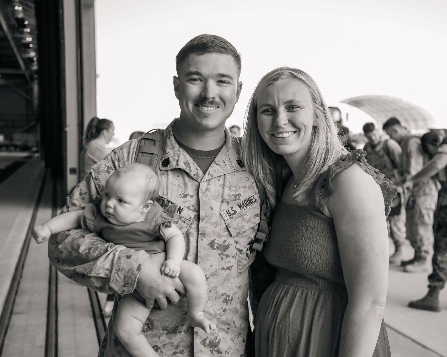 A series of five black and white photos showing emotional military homecoming moments with signs and American flags.