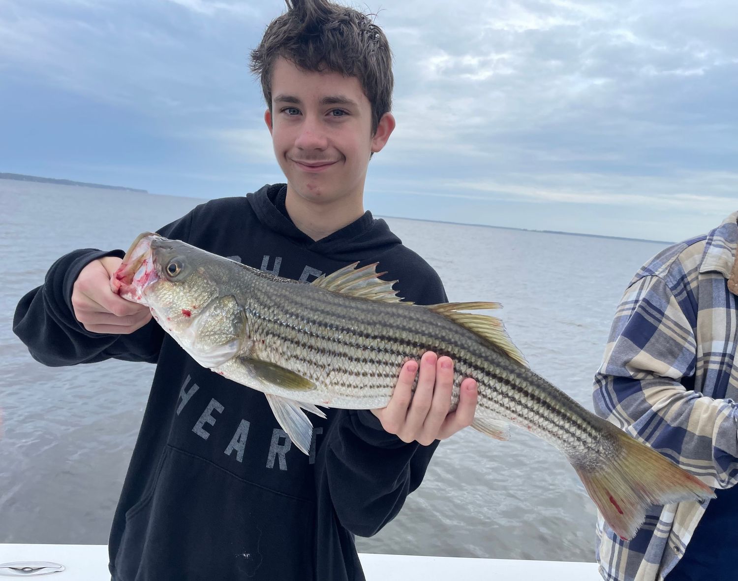 A striped bass catch being displayed near the water on an overcast day.
