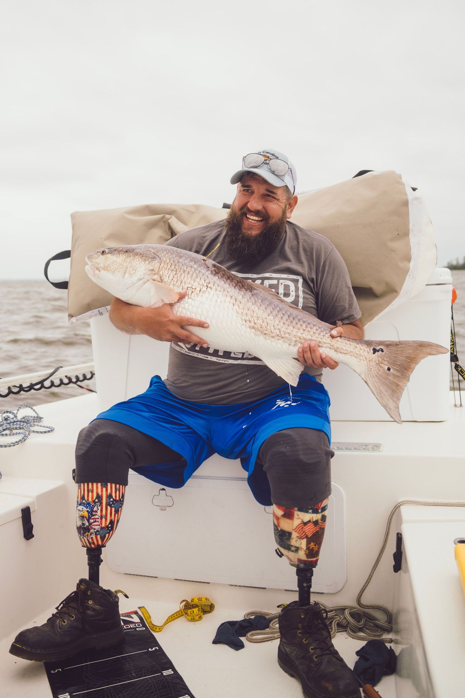 Smiling bearded man with prosthetic legs proudly holds a large redfish on a boat.