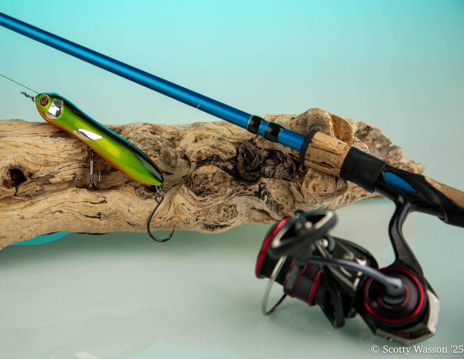 Blue fishing rod and reel resting on driftwood beside a green fishing lure with hooks on a light blue background.