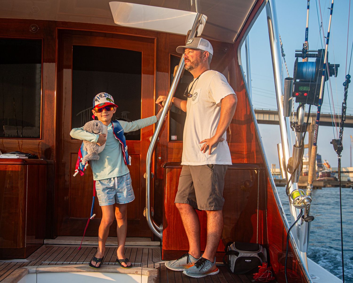 A child and adult fishing on a wooden sailboat deck at golden hour near a marina.