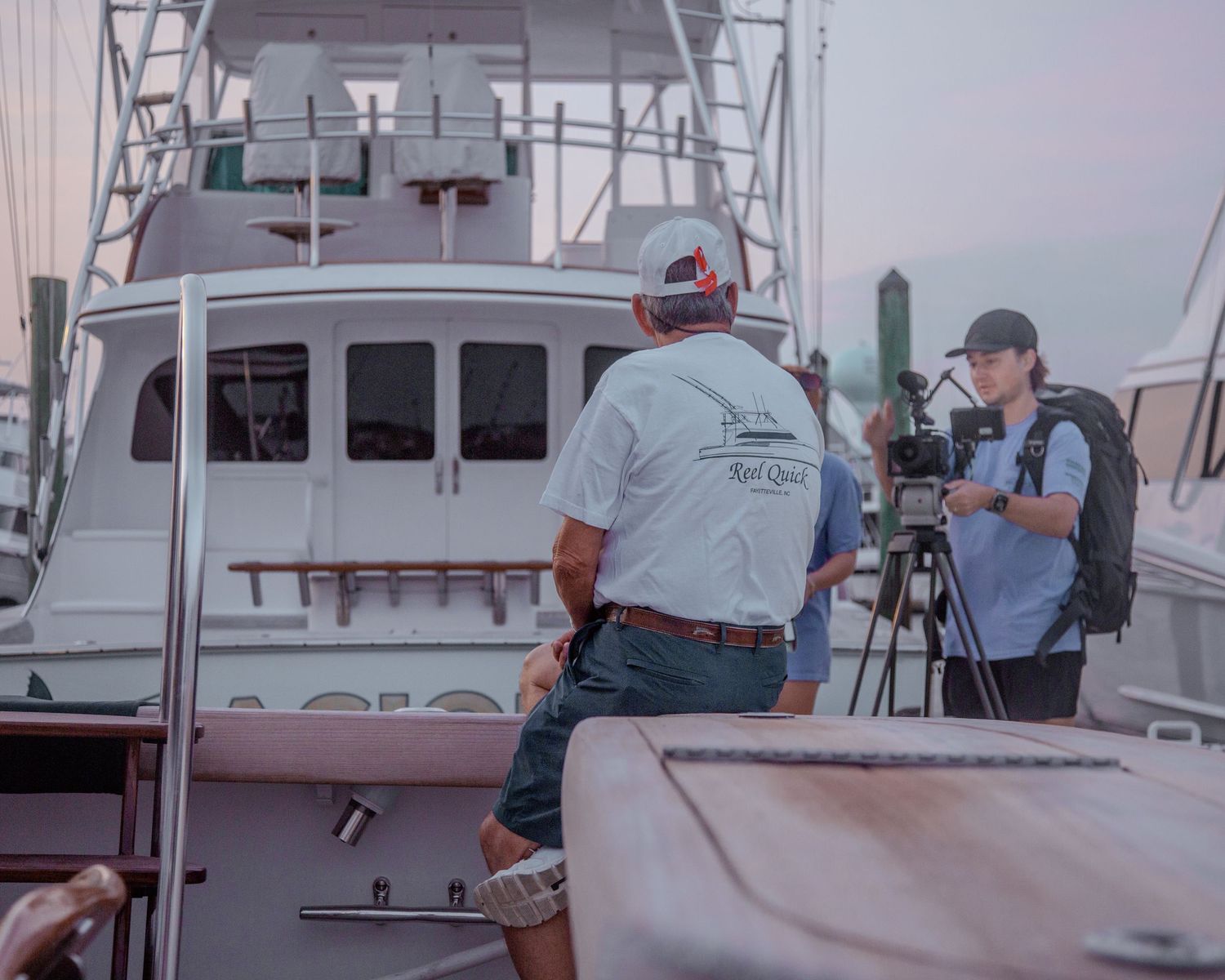 Two men at a marina dock, one wearing a Real Quest shirt near a large fishing boat while another films with a camera.