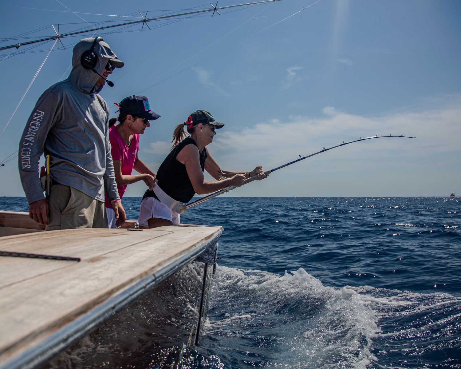 Three anglers fishing from a boat deck on deep blue ocean waters under a clear sky, woman bending rod with catch.