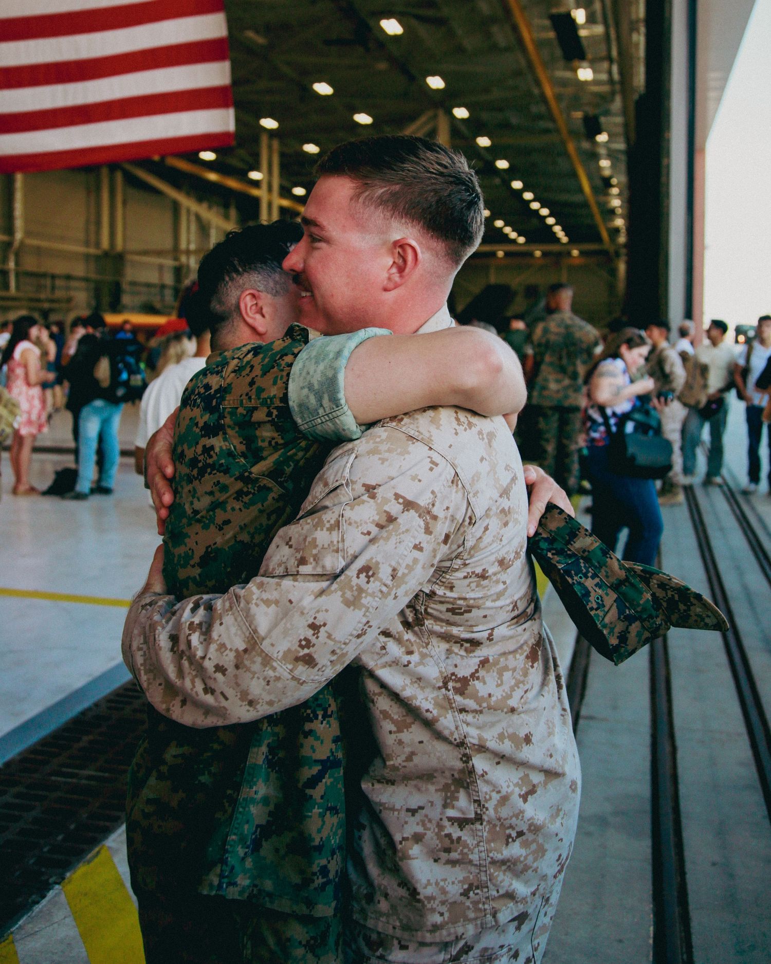 Military service members share emotional embraces during a homecoming ceremony in a hangar with American flags.